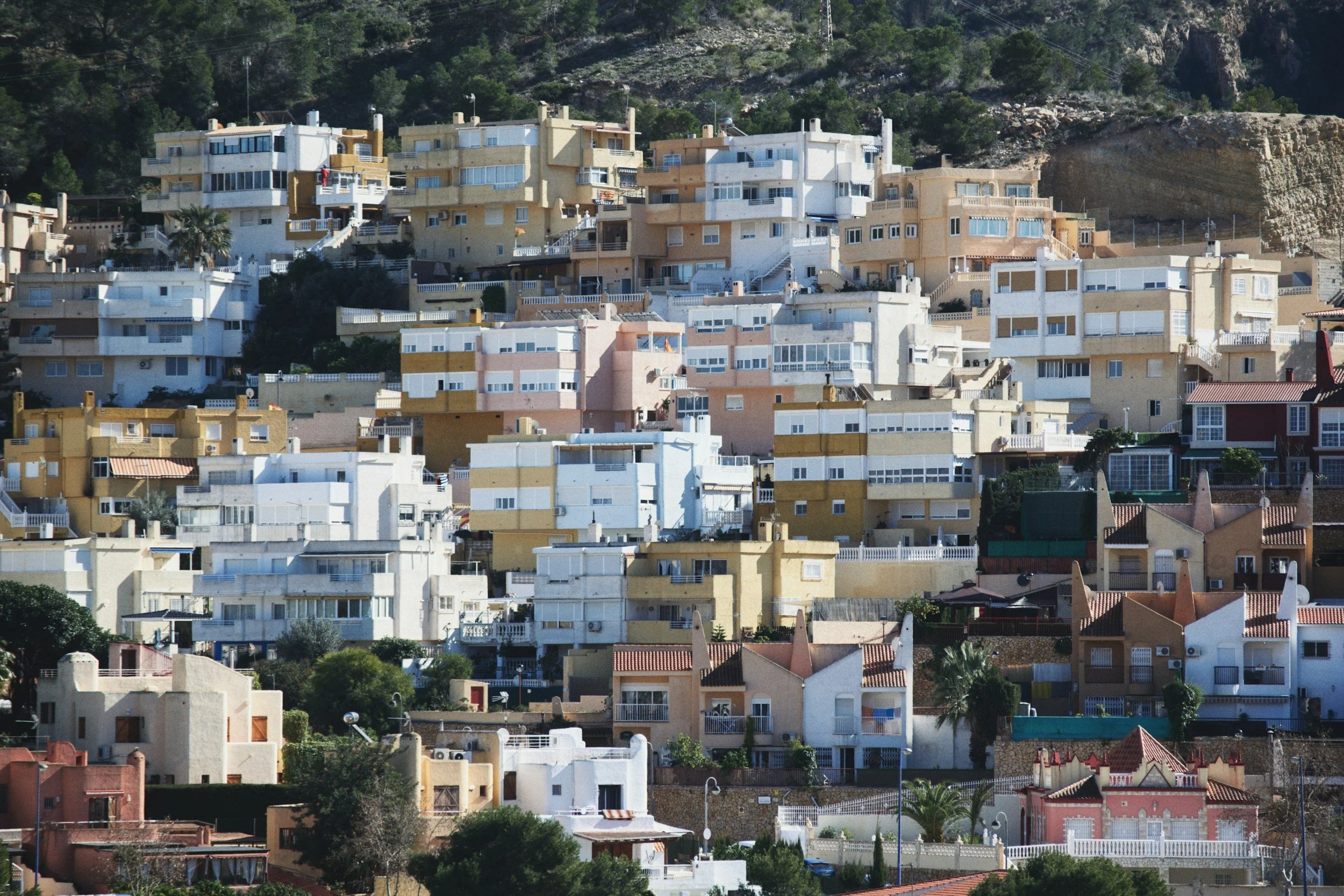 A hillside view of densely packed, multicolored residential houses, including white, cream, pink, and reddish-brown buildings with various roof styles, set against a backdrop of green trees and rocky terrain.