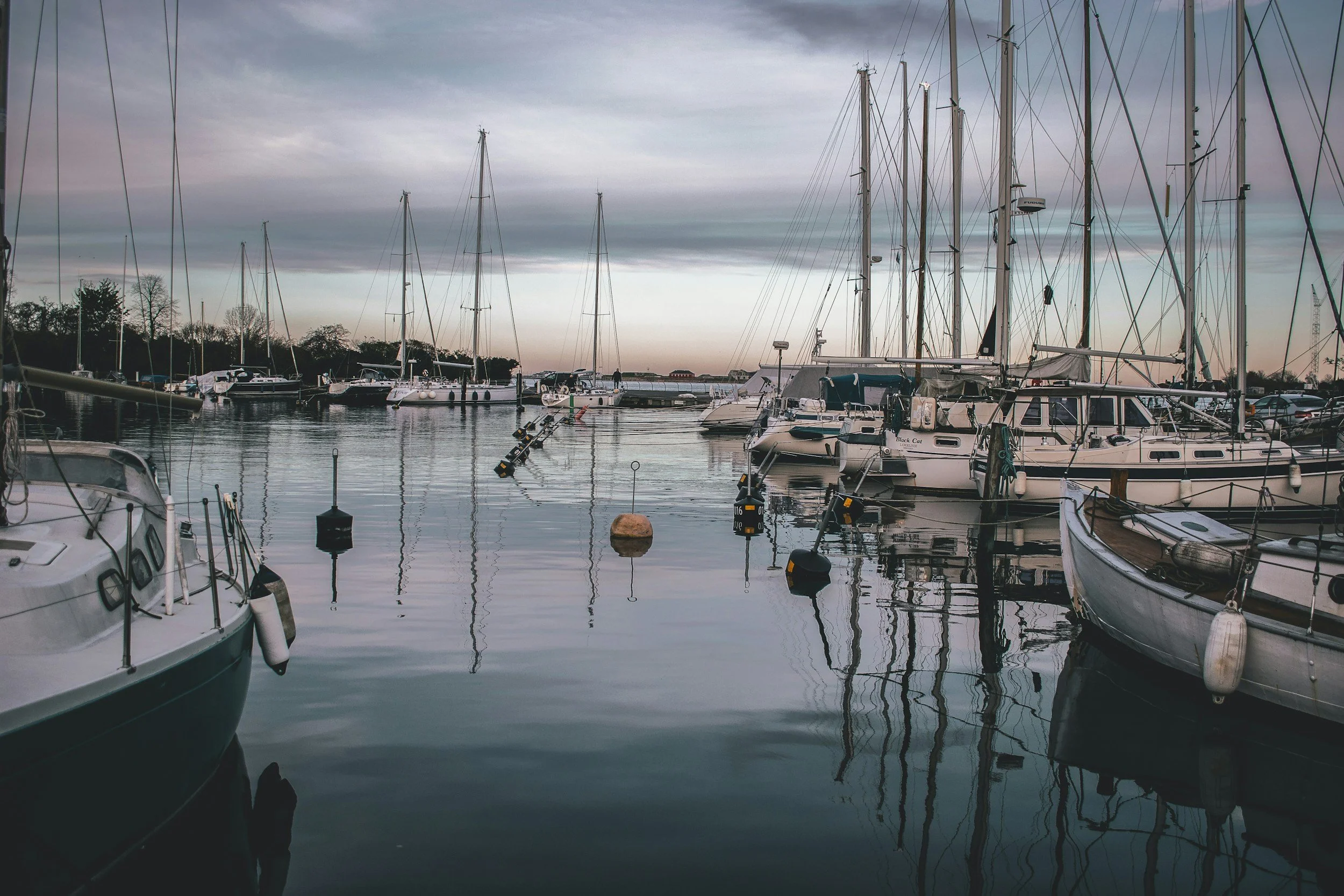 A marina at dusk with several sailboats and yachts moored, their reflections visible in the calm water, under a cloudy sky.