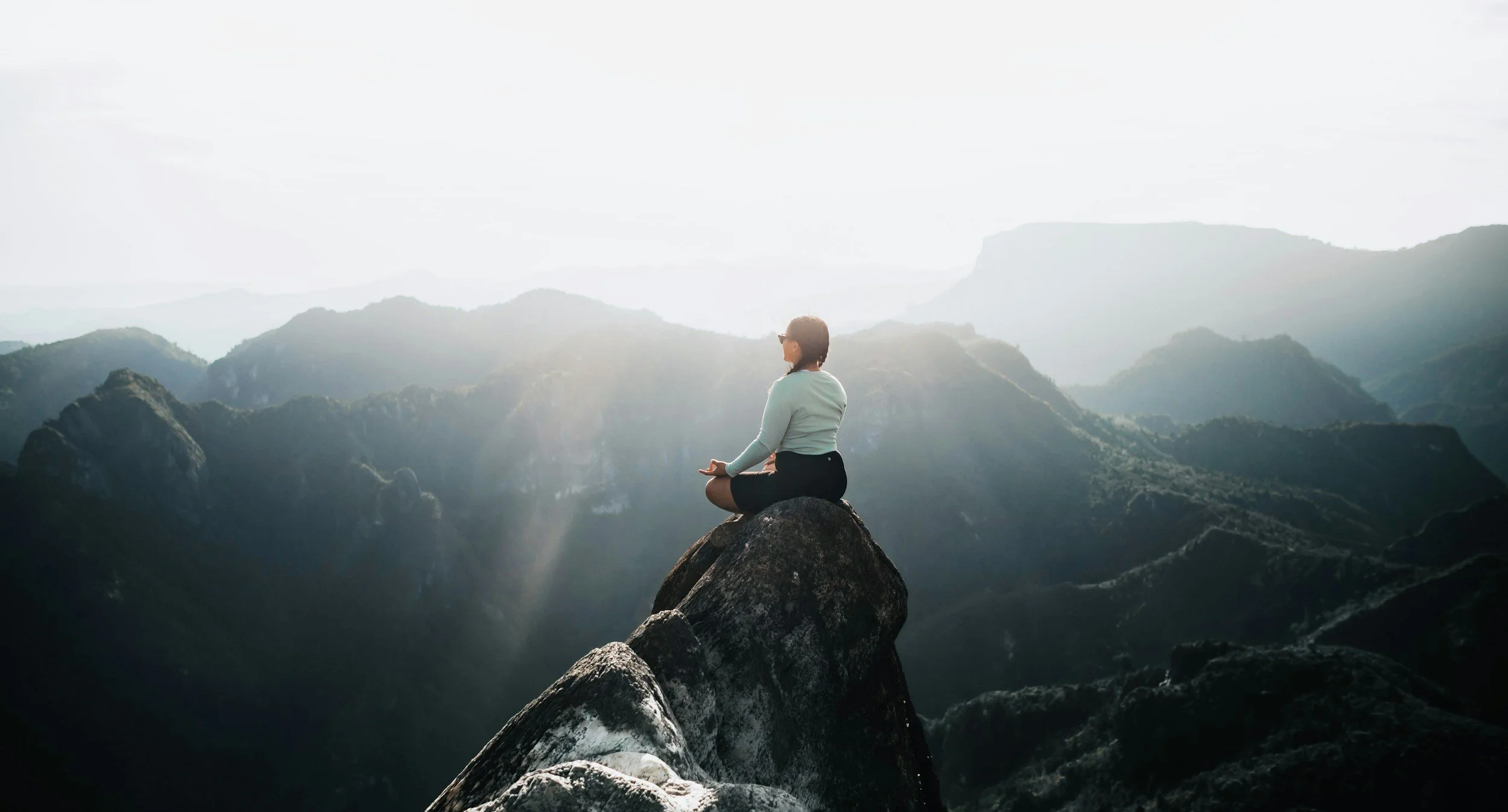 A woman practicing meditation or yoga while sitting cross-legged on a large rock amidst mountains with misty background, during sunrise or sunset.