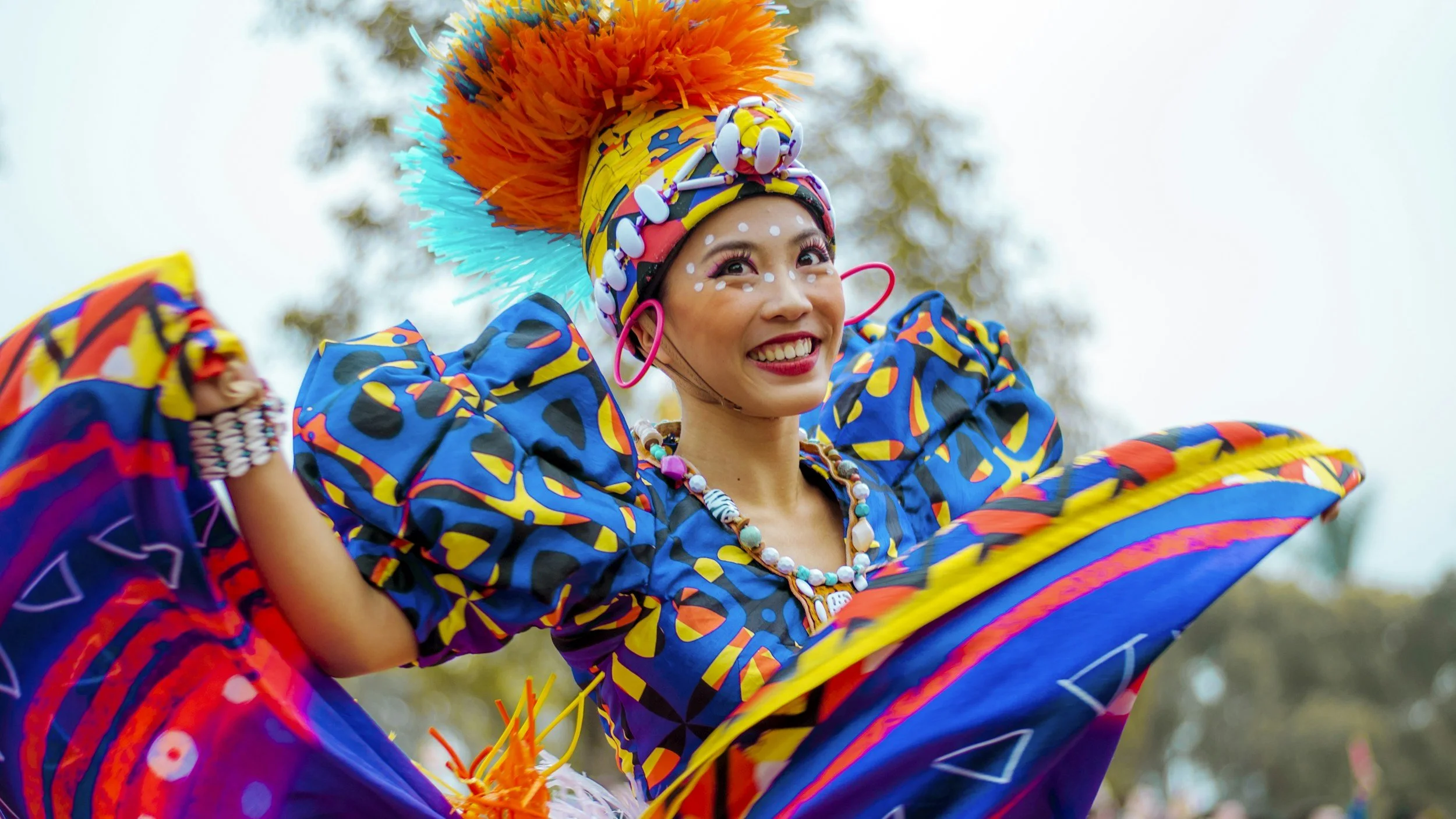 Woman in colorful traditional costume smiling outdoors, wearing a yellow headscarf with pom-poms and a dress with bright blue, orange, and yellow patterns, and large pink earrings.