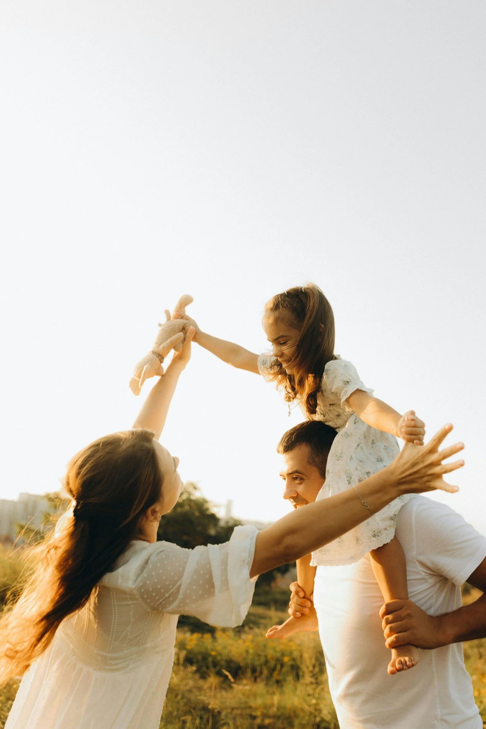 A family of four outdoors during sunset, playing and smiling, with a girl sitting on her father's shoulders and another girl reaching out to her mother.
