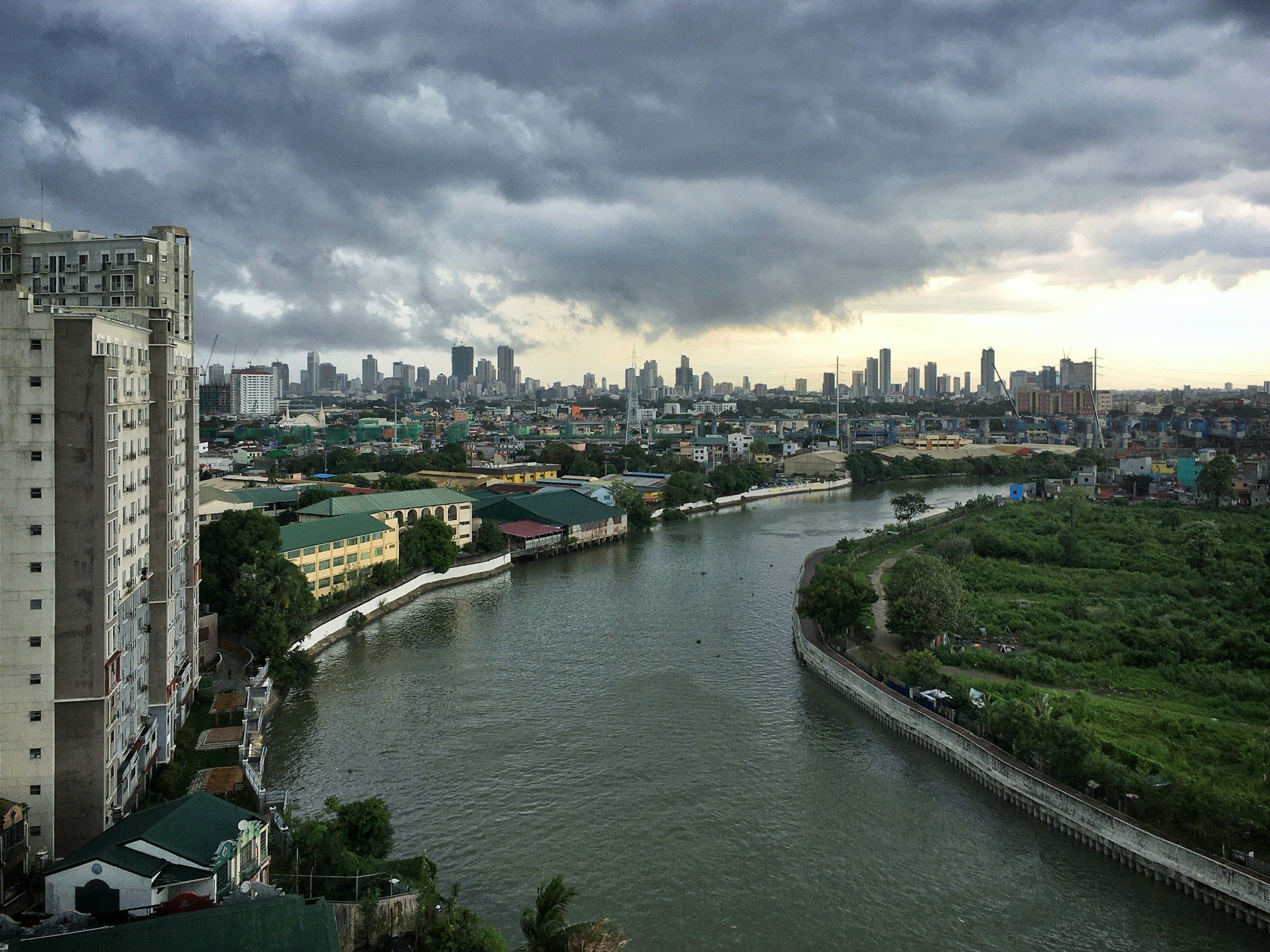 A city skyline with tall buildings and skyscrapers under a dark, cloudy sky, with a river and some greenery in the foreground.