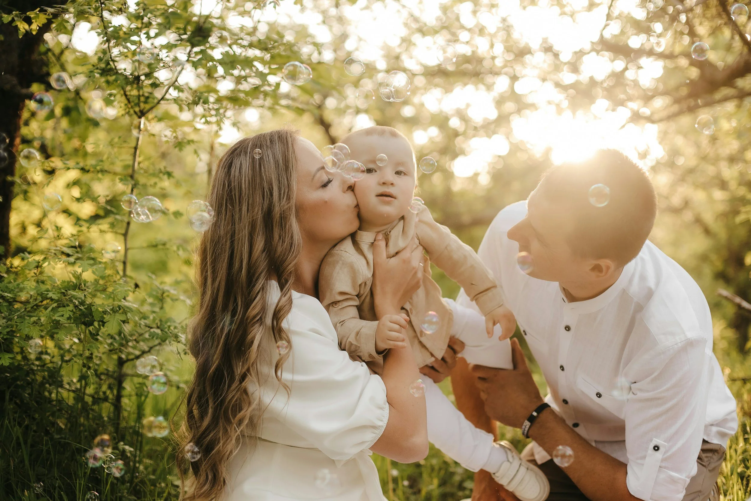 A woman with long hair holding a young boy with short light hair, while another person with short dark hair leans in to kiss the boy on the cheek. All are outdoors with sunlight filtering through trees, surrounded by green foliage and floating bubbles.