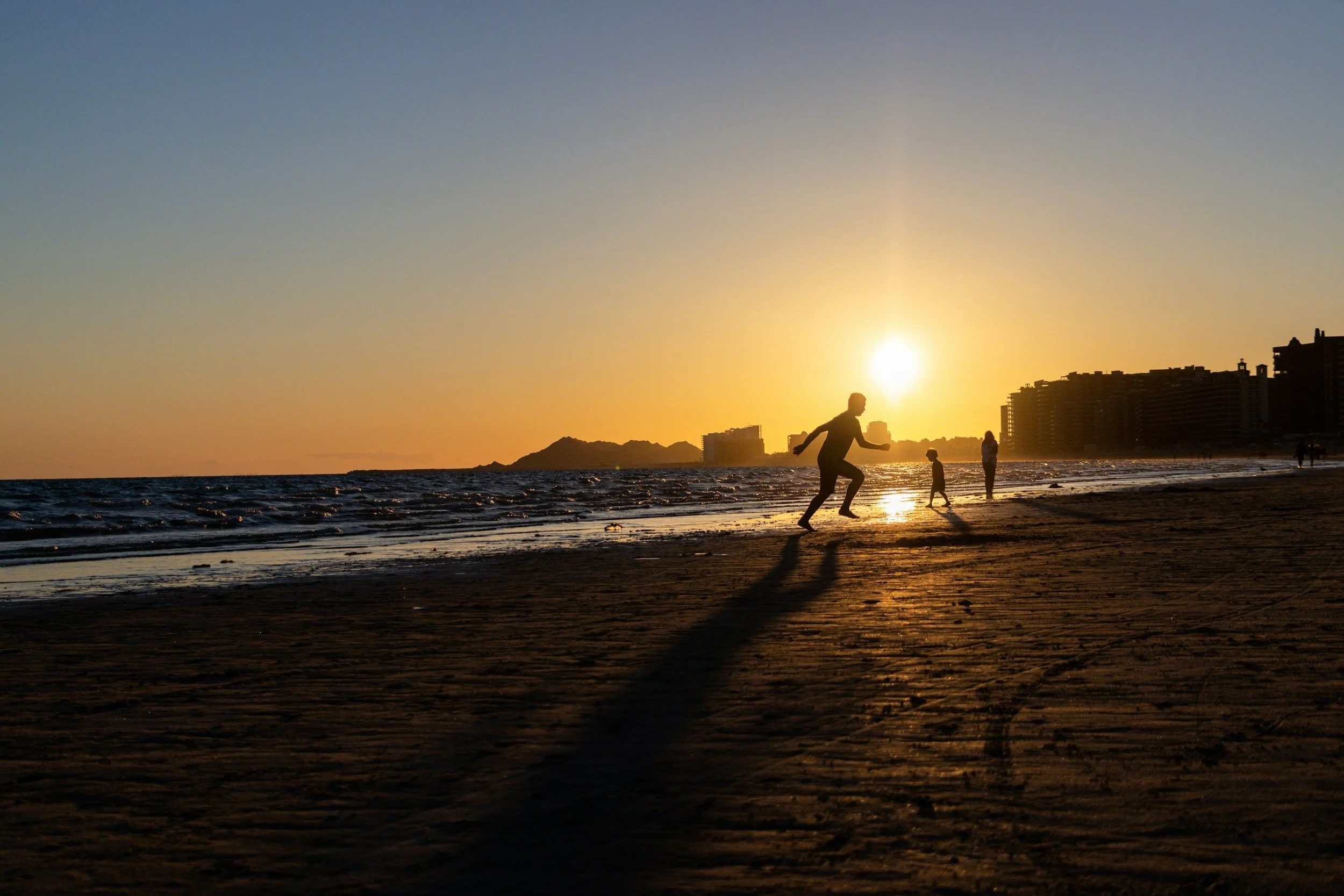 People playing on the beach during sunset with city buildings and mountains in the background.