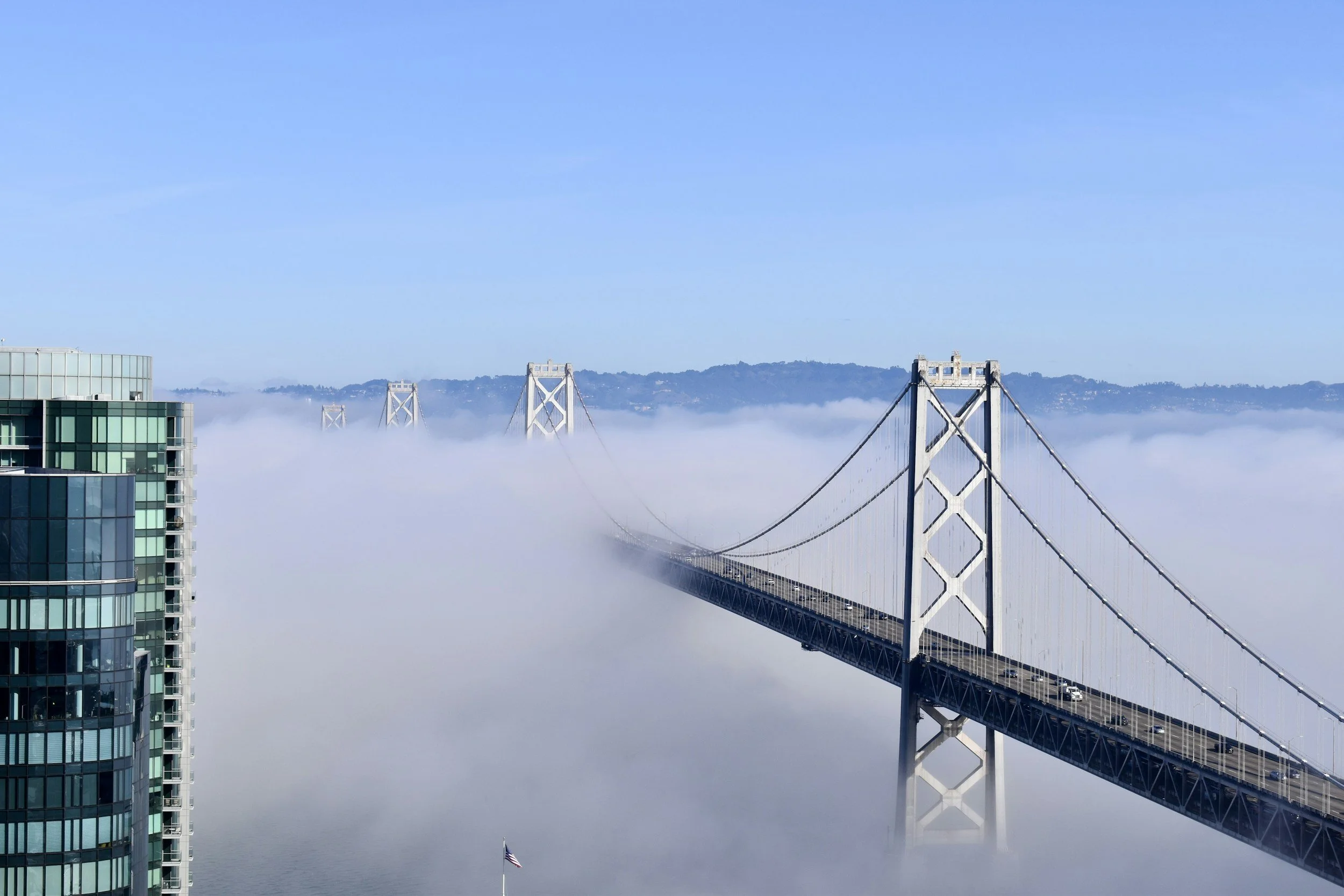 A bridge extending over foggy water with a city building on the left and a flagpole with an American flag below the bridge.