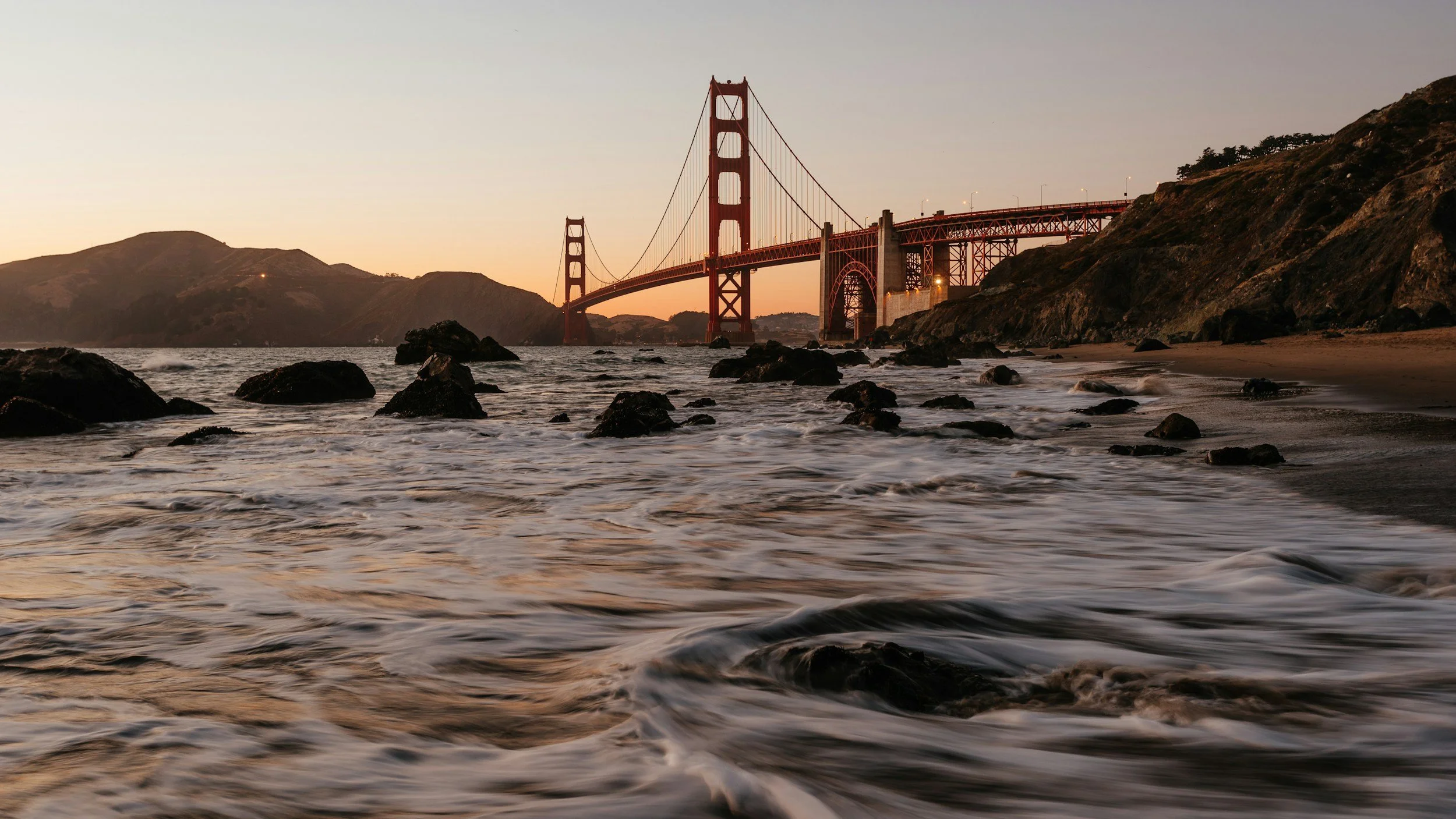 The Golden Gate Bridge in San Francisco at sunset, viewed from the shoreline with rocks and waves in the foreground.