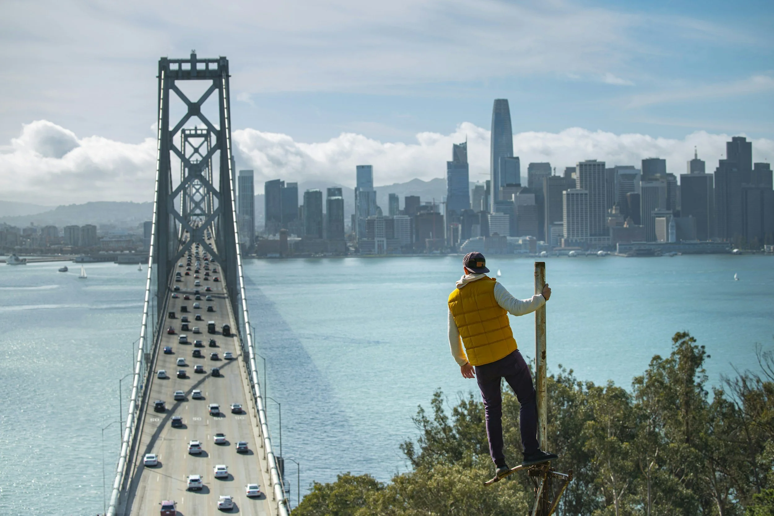 A person wearing a yellow vest and a baseball cap is riding a bicycle next to a railing overlooking a bridge and a city skyline, with water in the foreground and skyscrapers in the distance.