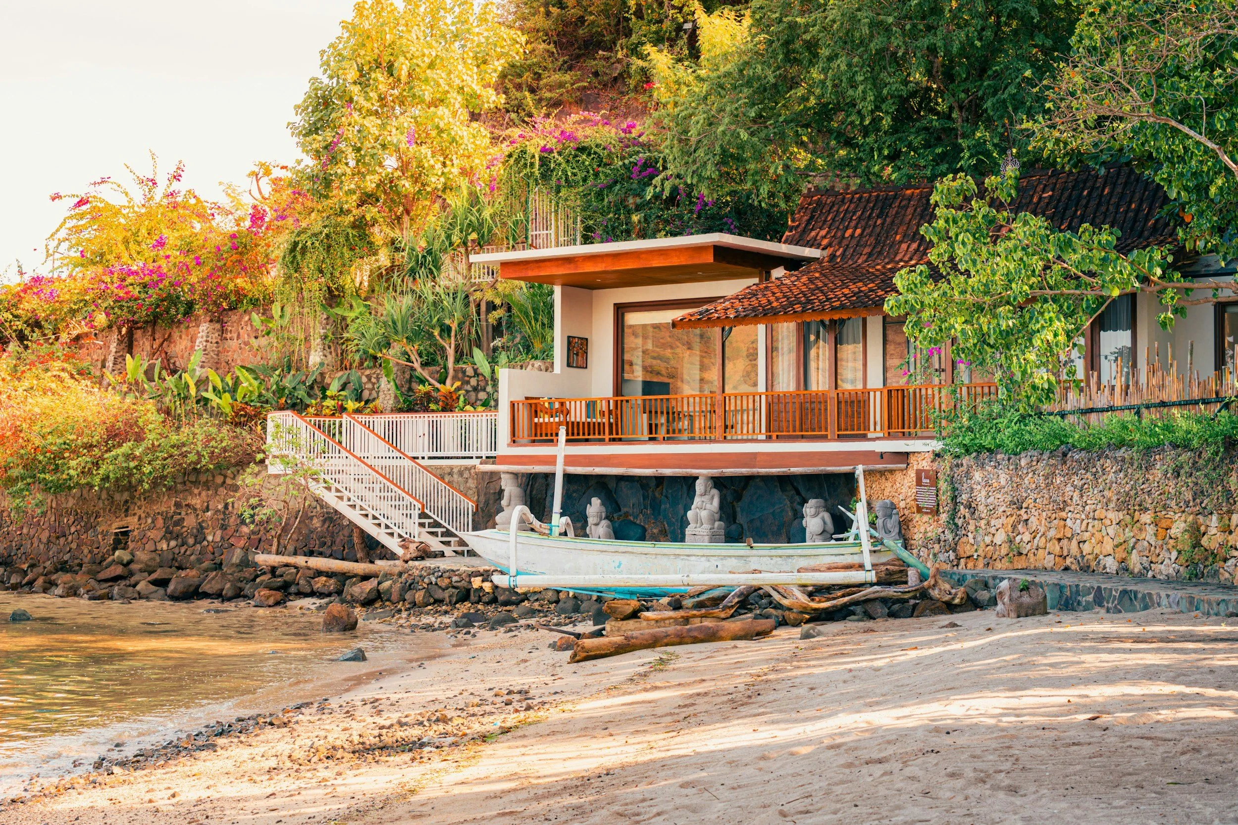 A house on a hillside with a balcony, surrounded by lush green trees and flowering plants, overlooking a body of water with rocks and driftwood on the shore.