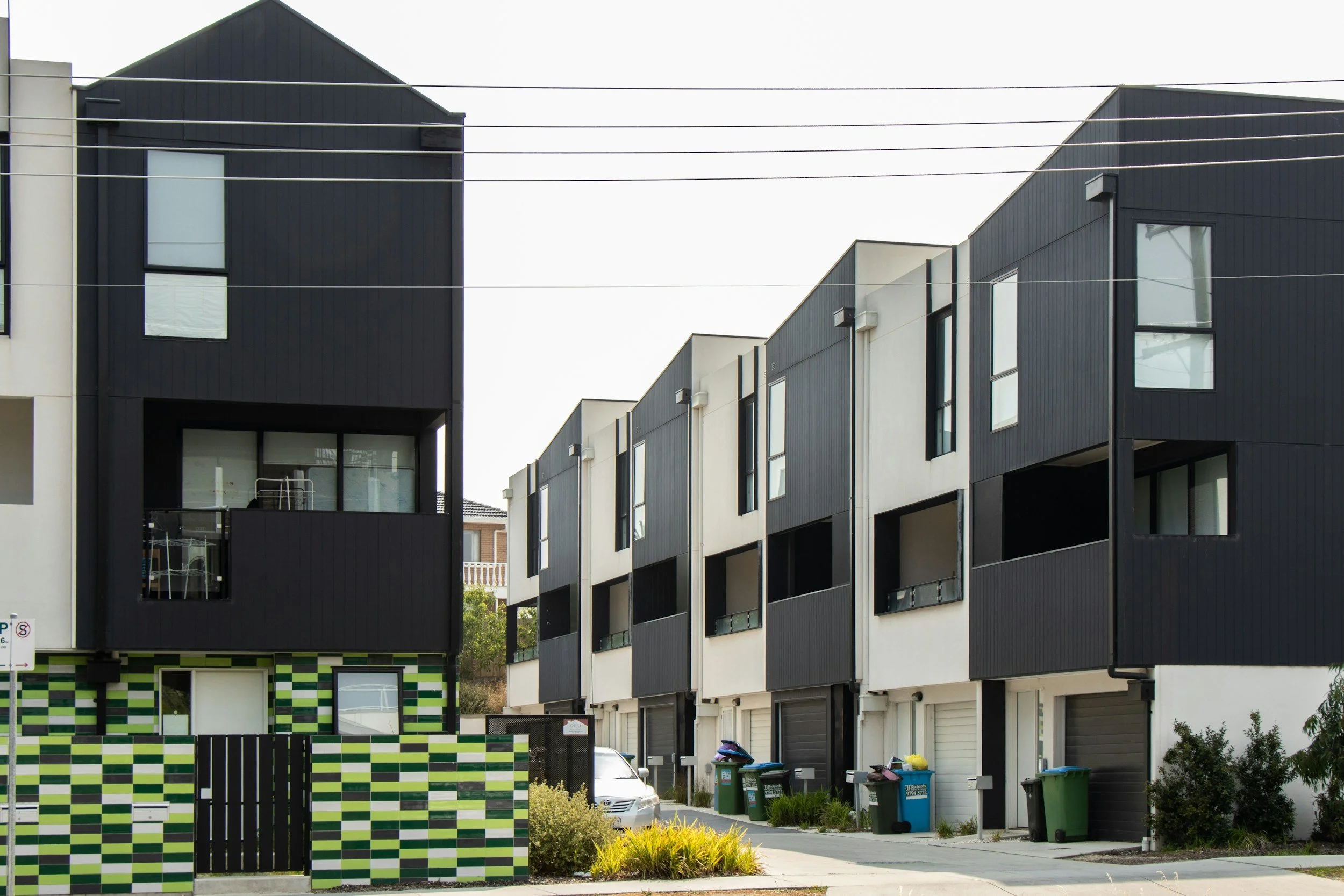 Modern row of black and white multi-story townhouses with large windows and garages, with waste bins and a parked car in front.