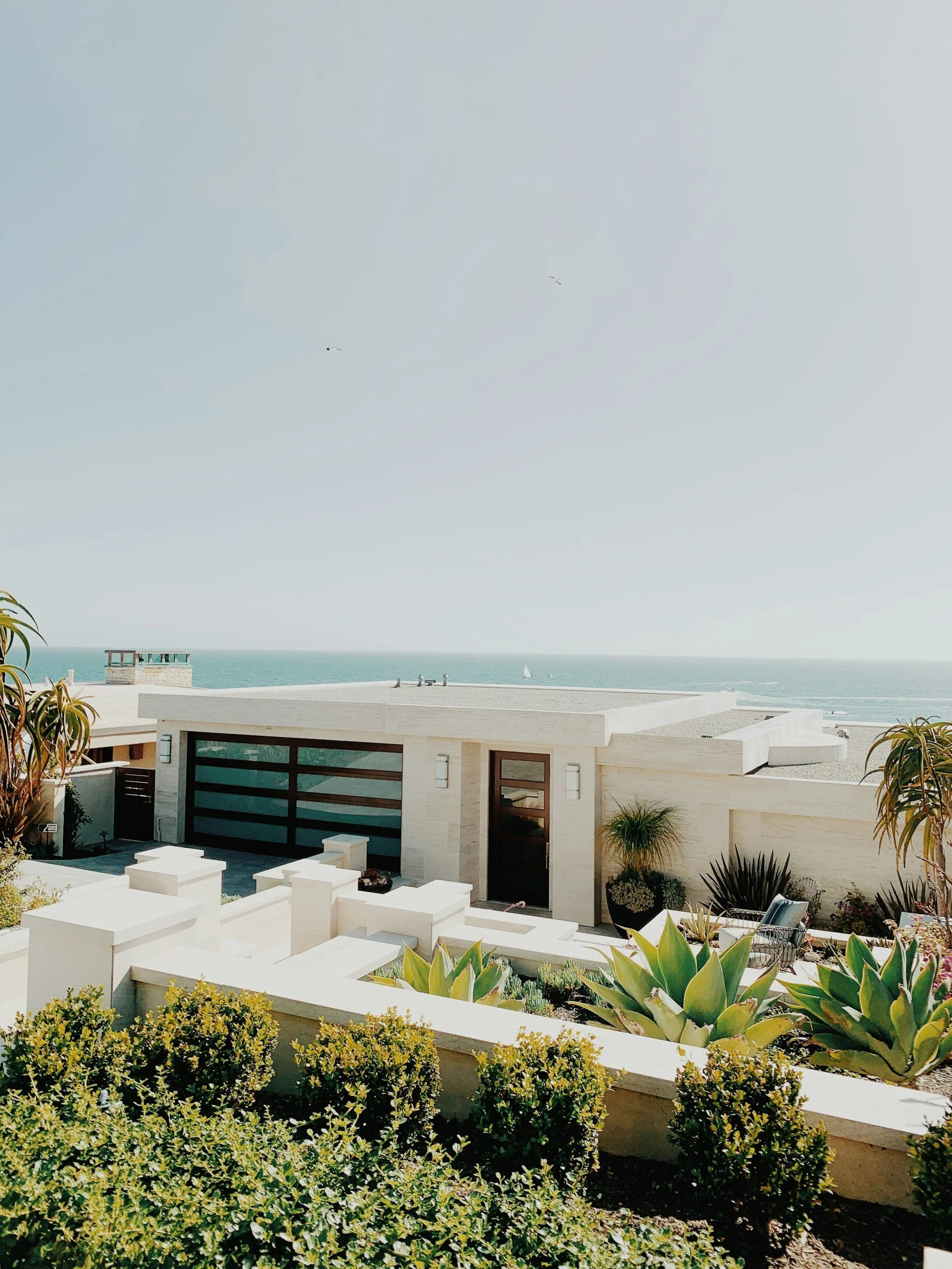 Modern white house with flat roof, large windows, and landscaped garden in front, overlooking ocean with sailboats in the distance under a clear sky.