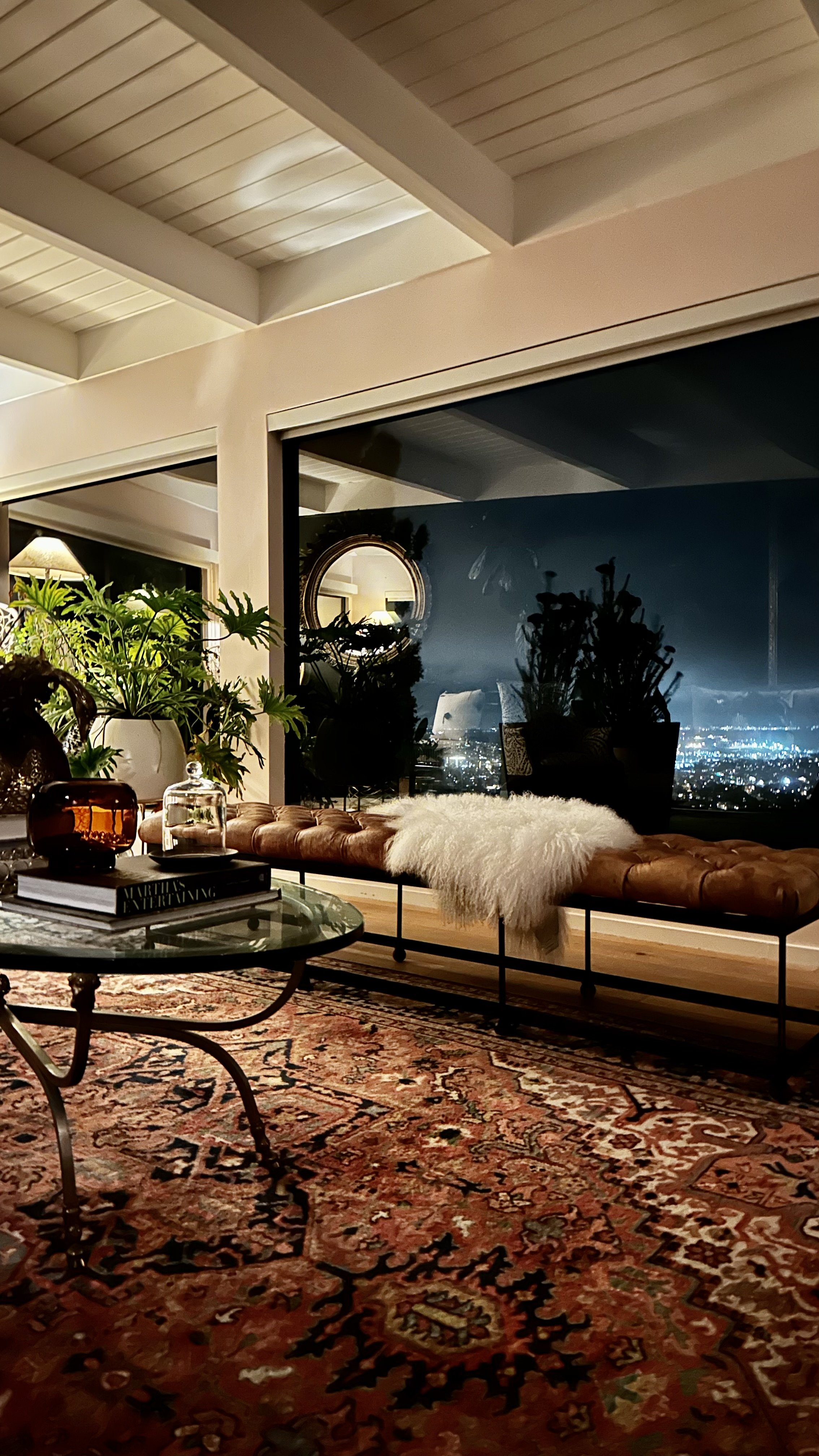 Living room with large window showing a cityscape at night, decorated with plants, a tan leather bench with a white fur throw, a glass coffee table with books and decorative items, a colorful patterned rug, and a mirror on the wall.
