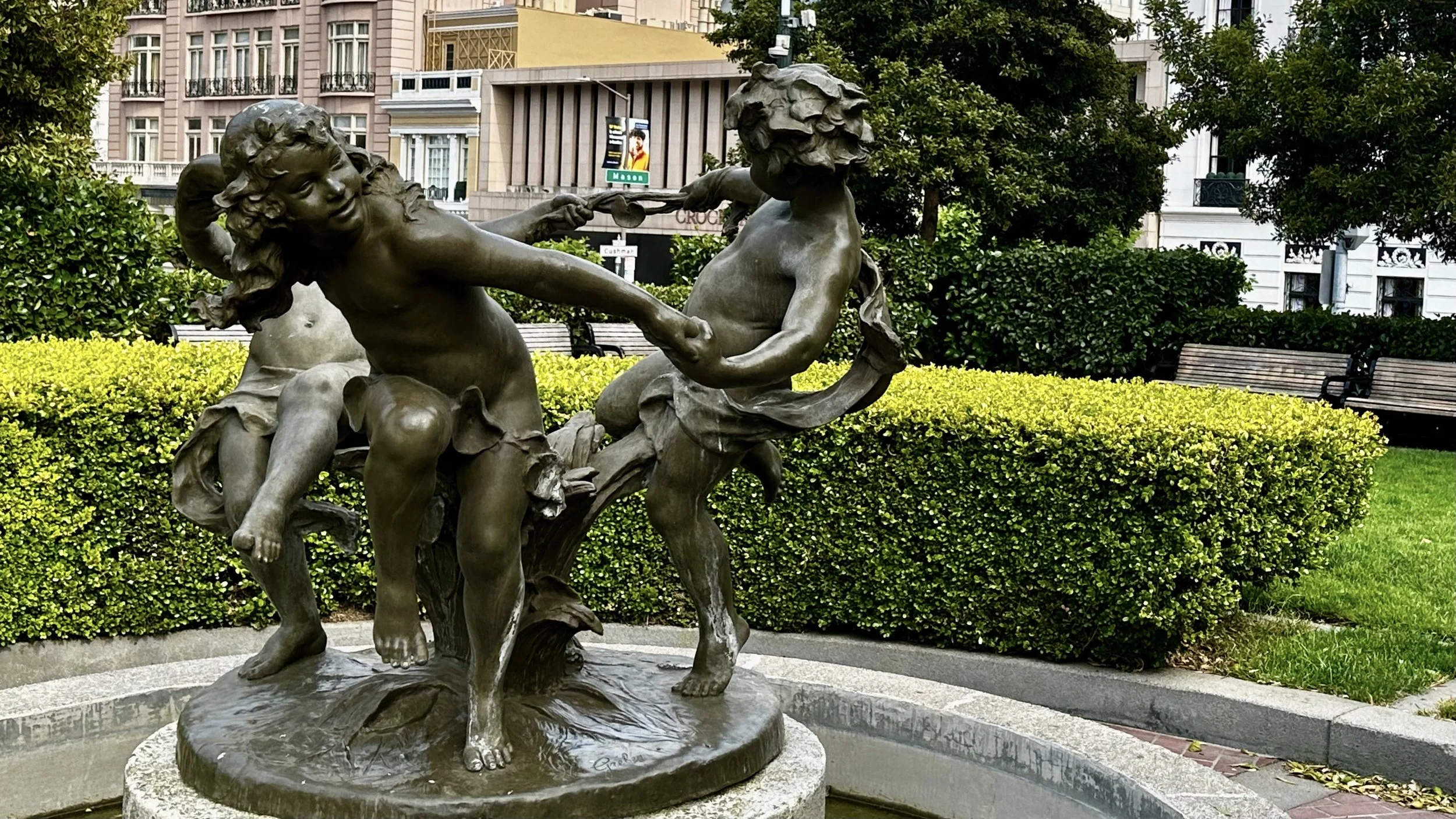 Bronze sculpture of two children playing tug-of-war, set in a park with green bushes, benches, and buildings in the background.