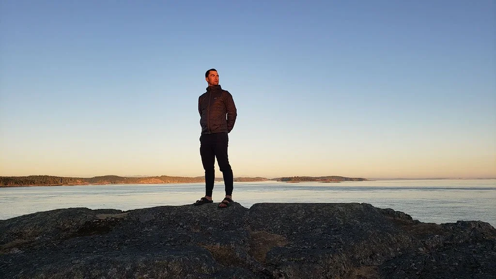 A man standing on a large rock near the ocean during sunset, with a clear sky and distant shoreline.