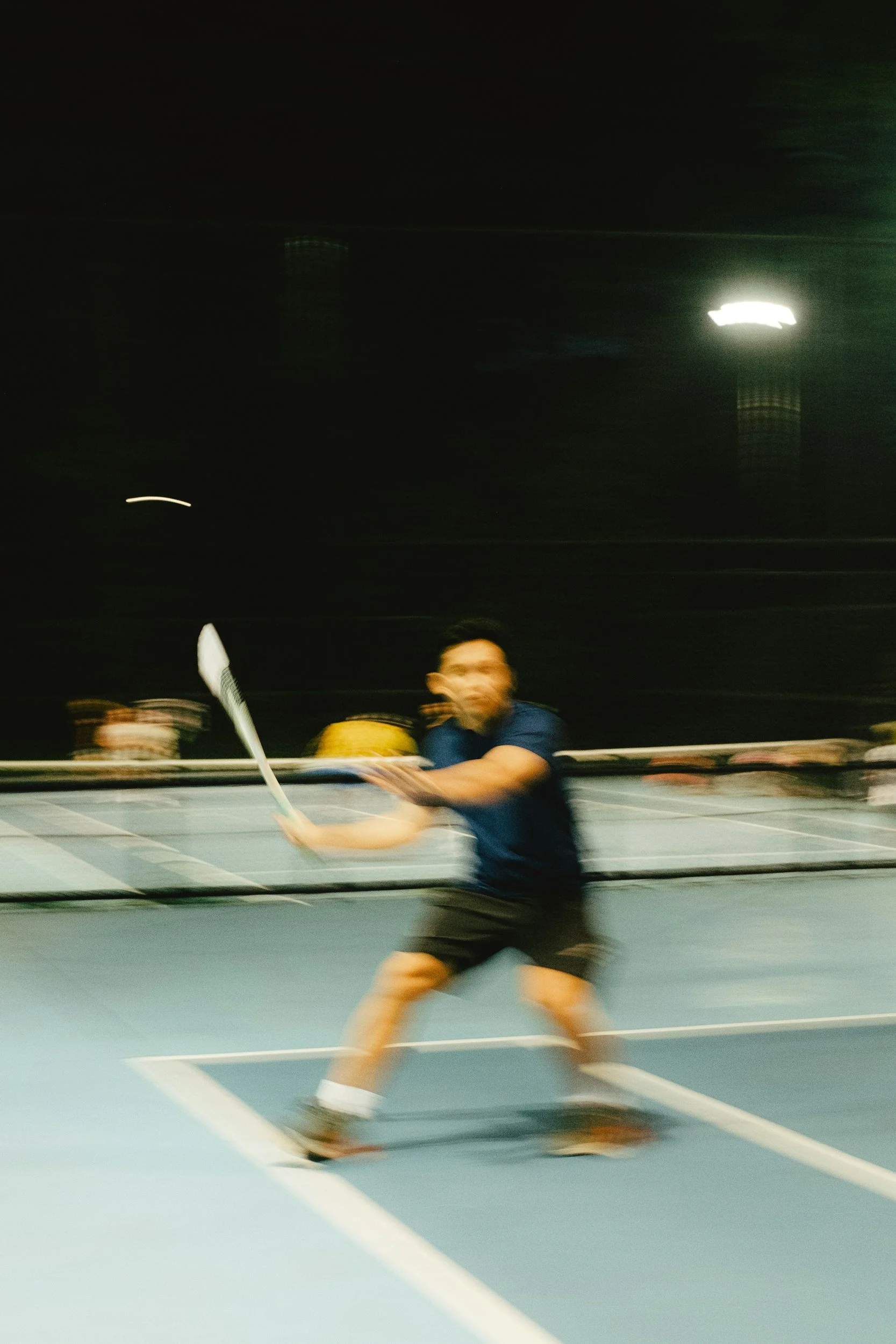 A person playing tennis on an indoor court, holding a tennis racket, with motion blur indicating movement.