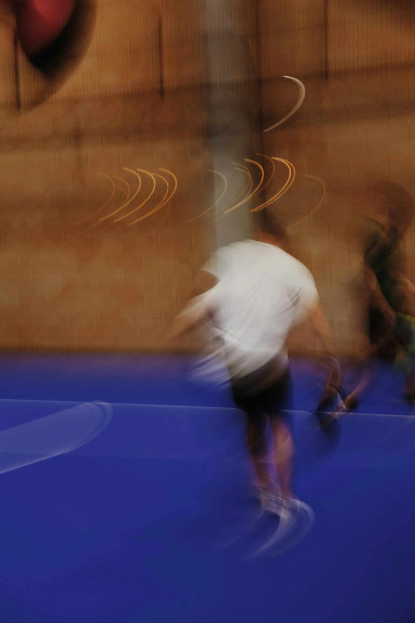 Blurred image of a person in a white shirt and black shorts playing indoor soccer on a blue court.