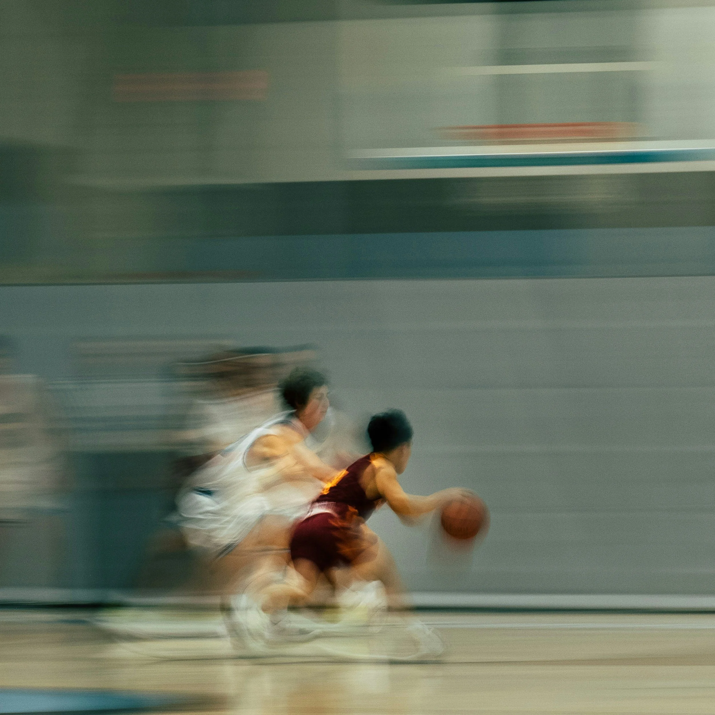 Blurry action shot of two young boys playing basketball on an indoor court, one in a white jersey and the other in a maroon jersey, dribbling the ball.