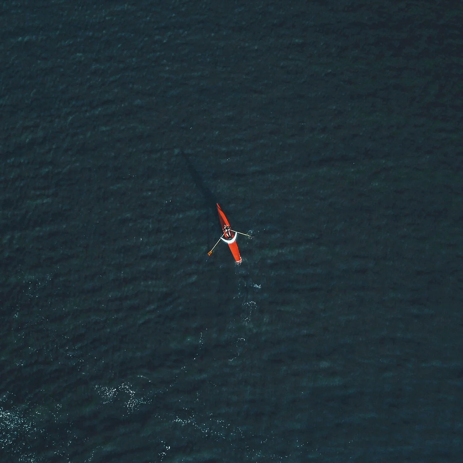 An aerial view of a red sailboat sailing on dark blue water, creating ripples around it.