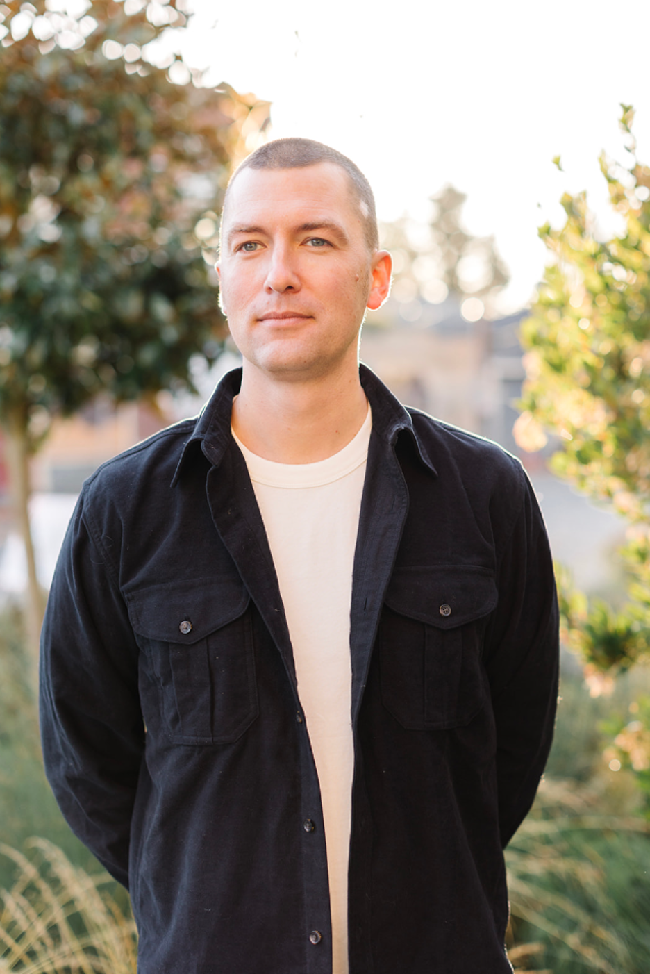 A young man stands outdoors during daytime with trees and foliage in the background. He has short hair, wears a black jacket over a white t-shirt, and looks slightly to the right.