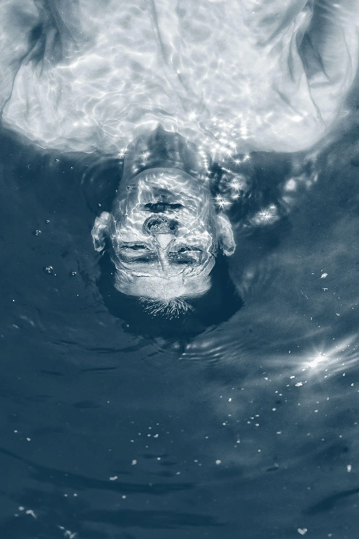 A person floats face up in water with their eyes closed and face partially submerged, creating ripples and sparkles on the water surface.
