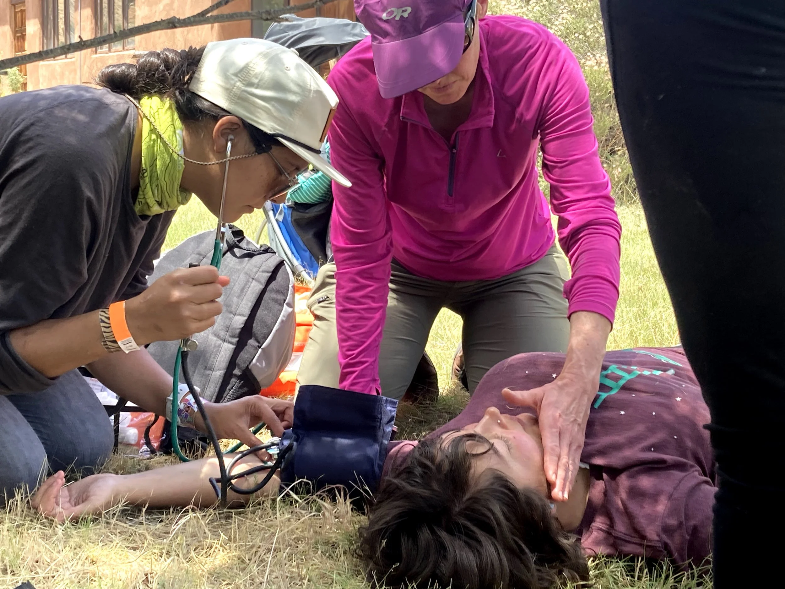 A first aid scenario with two people attending to someone lying on the ground. One person is using a stethoscope and blood pressure cuff, while the other appears to be checking the individual’s condition. They are outdoors on grass.