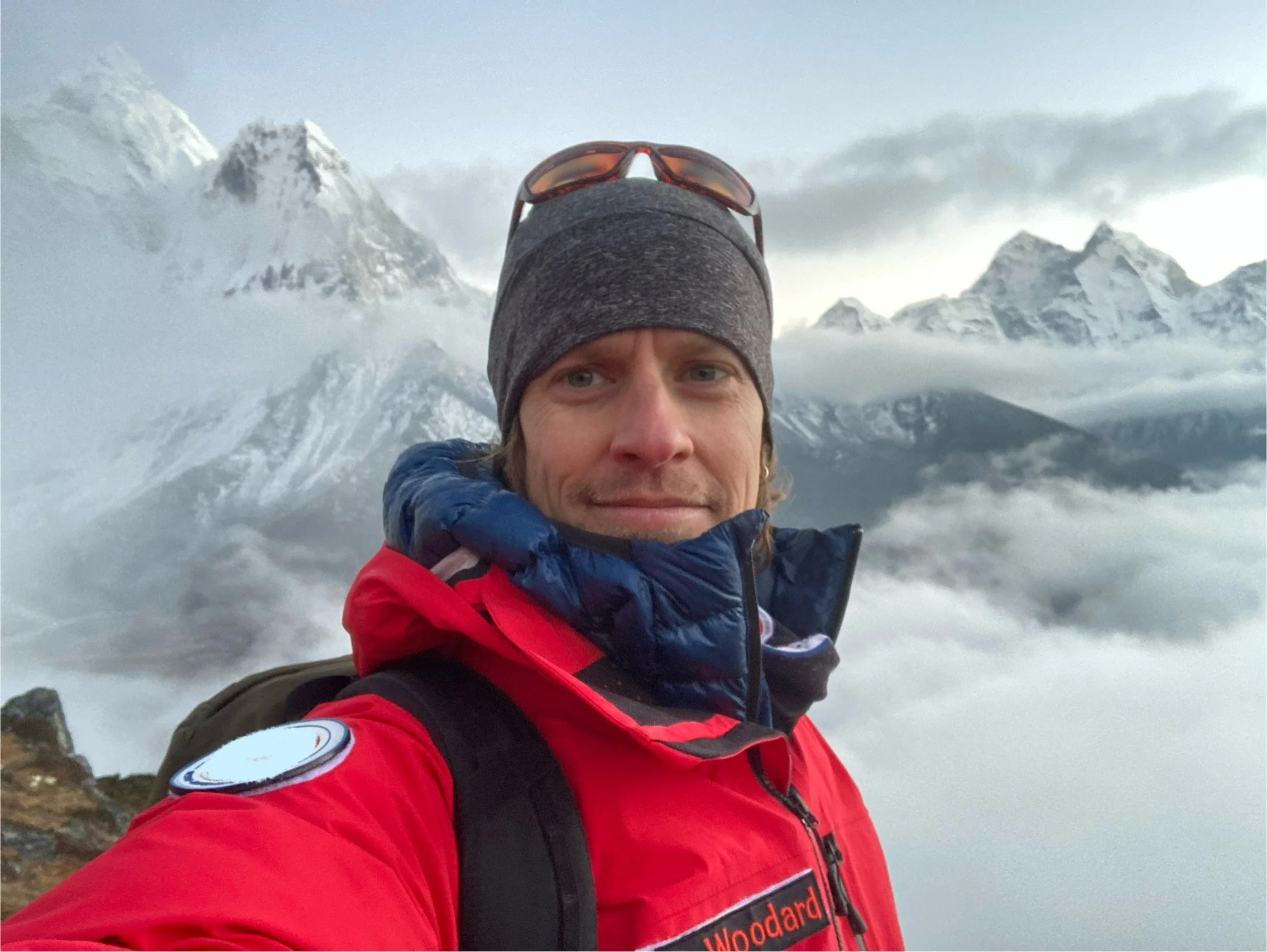 Person wearing a red jacket and beanie, standing in a mountainous landscape with snowy peaks and clouds in the background.