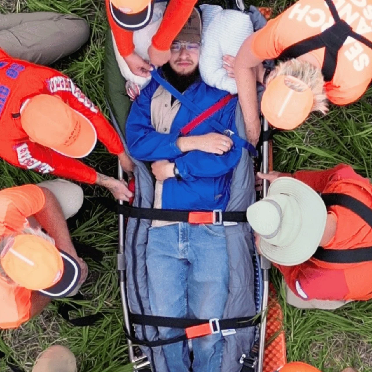 A group of search and rescue team members in orange uniforms assisting a person lying on a stretcher in a field.