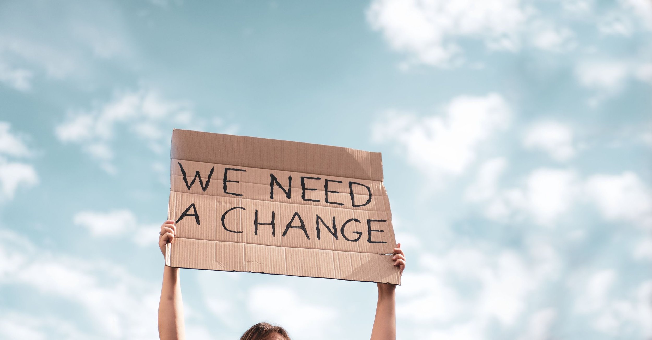 Person holding a cardboard sign that says 'We Need a Change' against a cloudy sky background.