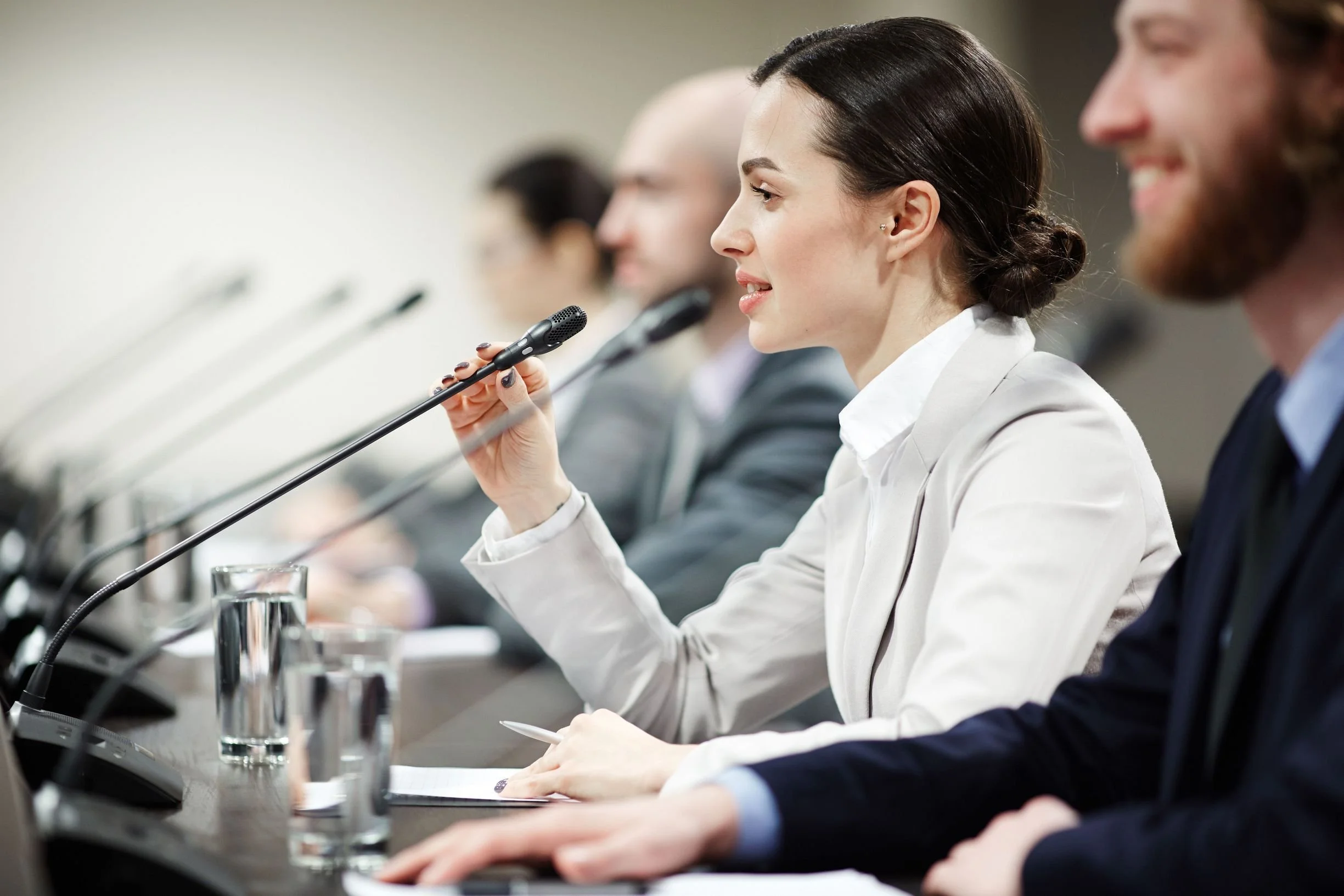 A woman in a white blazer speaking into a microphone at a conference or meeting.
