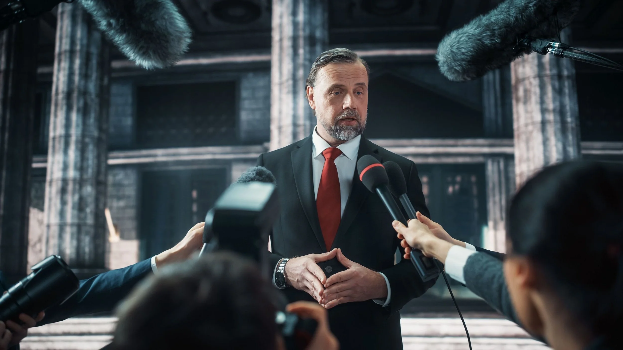 A middle-aged man with a beard and mustache, dressed in a black suit with a white shirt and red tie, speaking to multiple reporters holding microphones and recording devices, in front of a building with large columns.