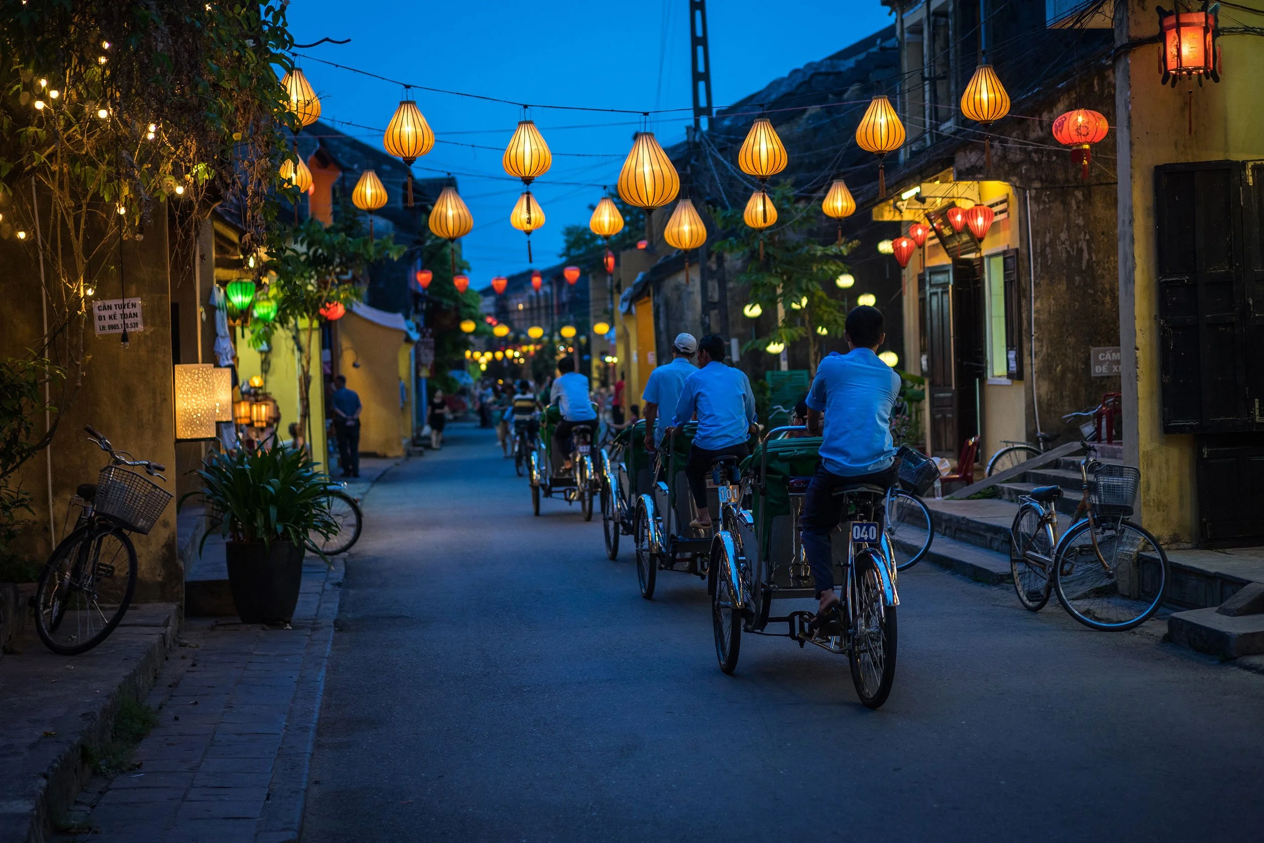 People riding bicycles on a street decorated with colorful lanterns during dusk in a lively neighborhood.