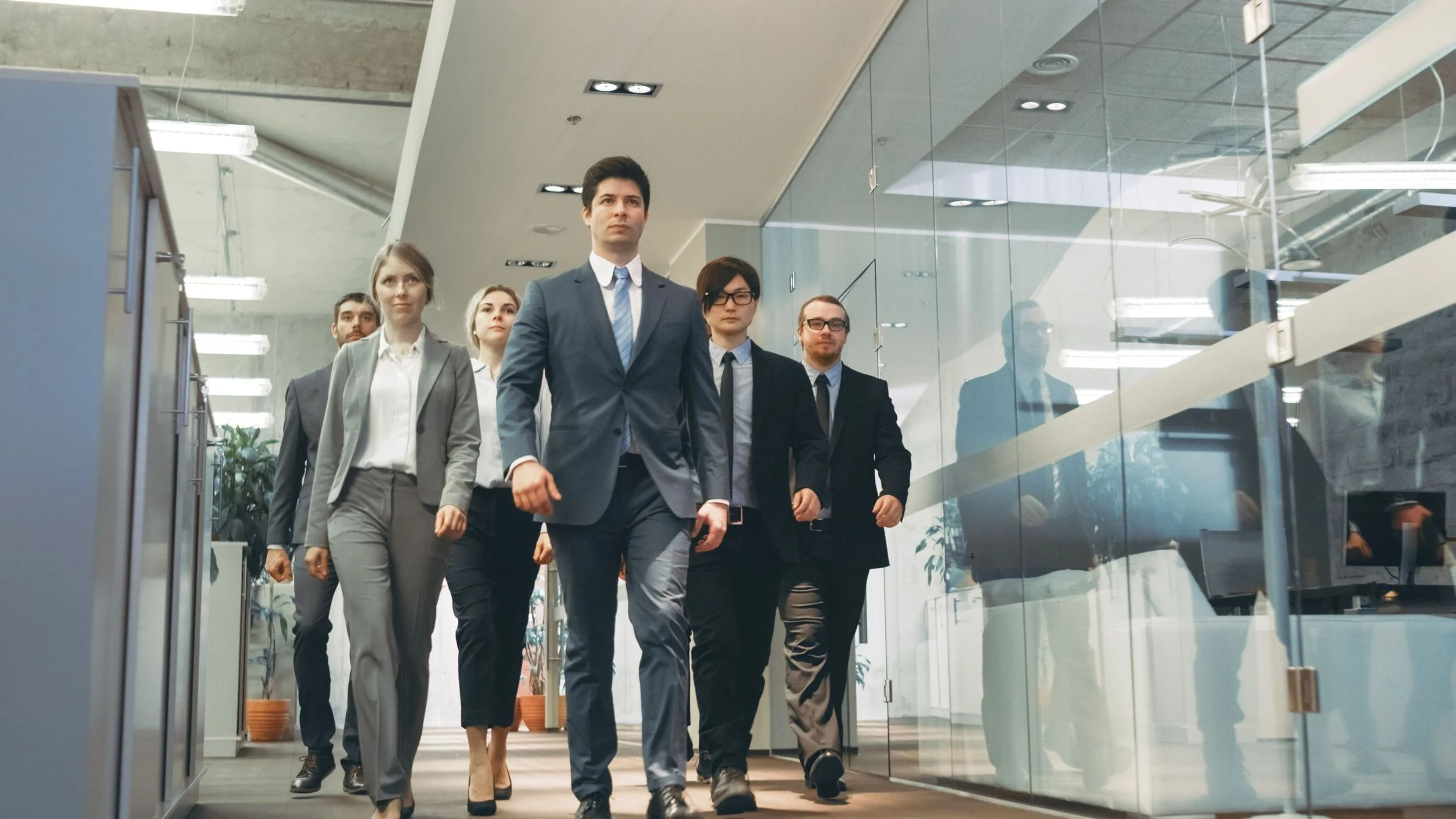 Group of six professionally dressed people walking through a modern office corridor with glass walls.