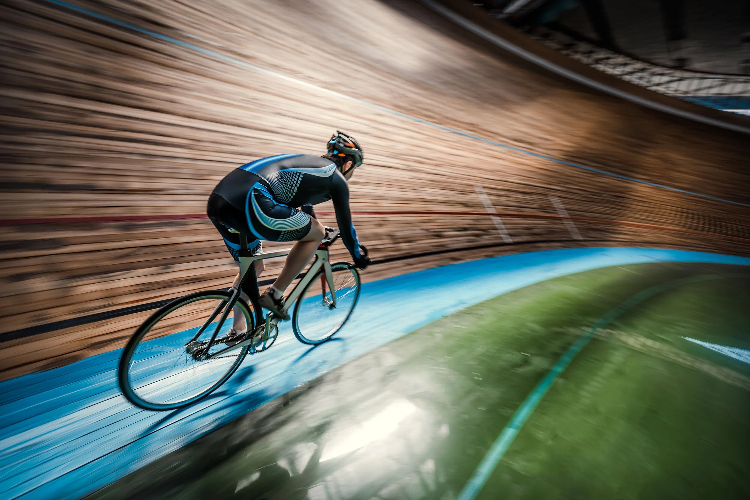 A cyclist dressed in black and blue gear riding on an indoor velodrome track with wooden walls, captured in motion.