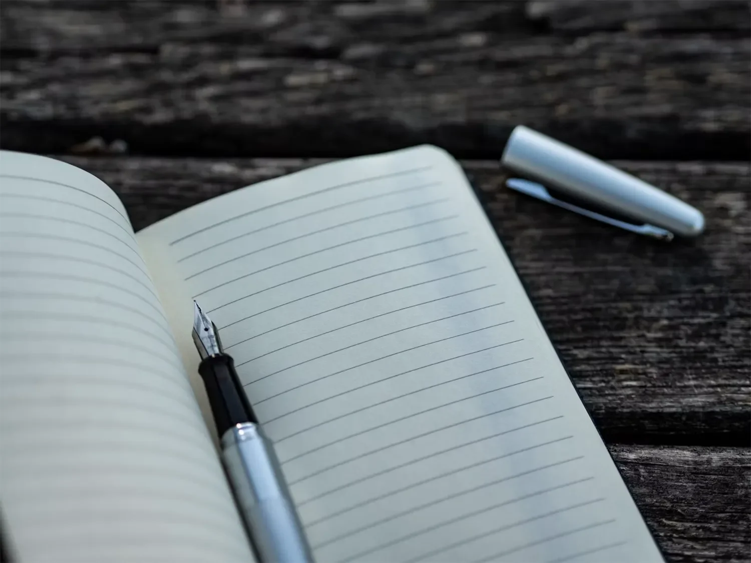 An open lined notebook with a pen resting on it, placed on a wooden surface, with a pen cap nearby.