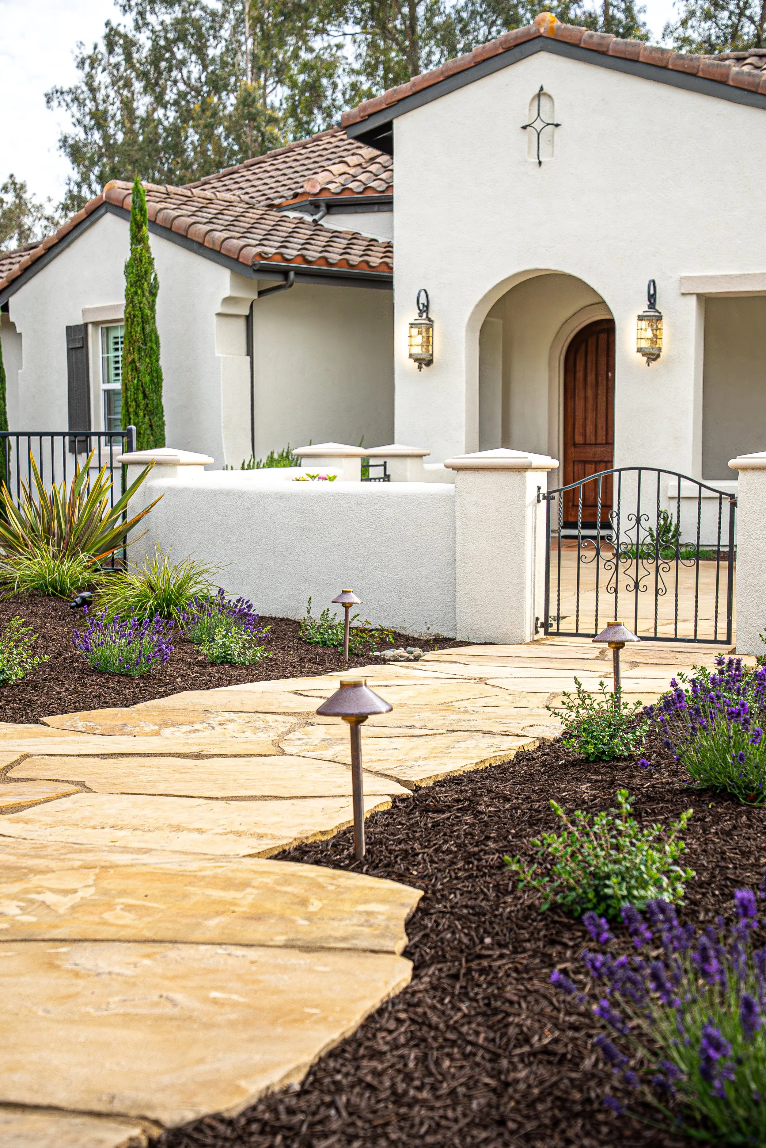 Front yard of a white house with a tiled roof, arched entryway, wooden front door, black wrought iron gate, landscaped garden with purple flowers, and decorative pathway lighting.