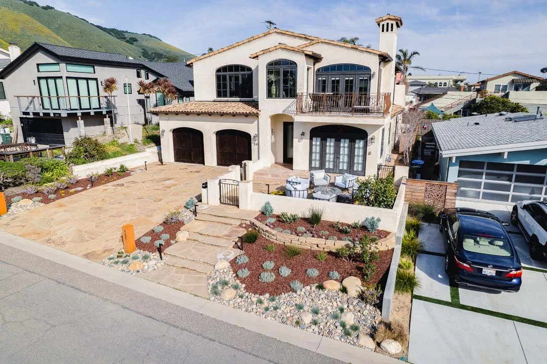 A two-story Mediterranean-style house with a tiled roof, large arched windows, and a balcony. Front yard with stone walkway, small garden, and patio furniture, and two garage doors. designed and built by Chaparral Pavers, the top high end landscaper