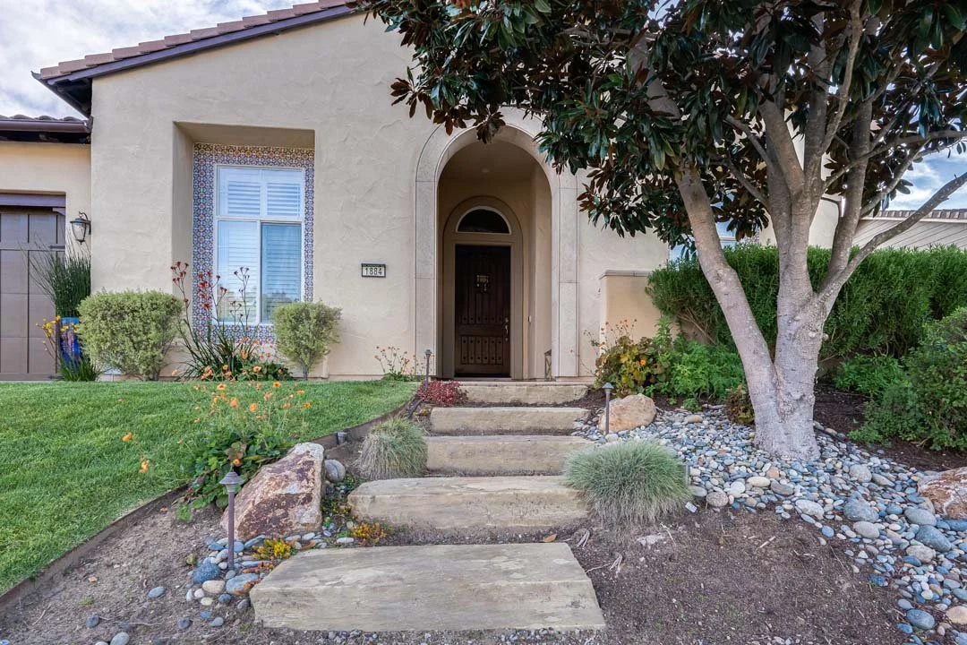 Front yard of a house with a pathway made of large stepping stones leading to a front door, surrounded by bushes, a tree, and landscaped garden with rocks and flowers.