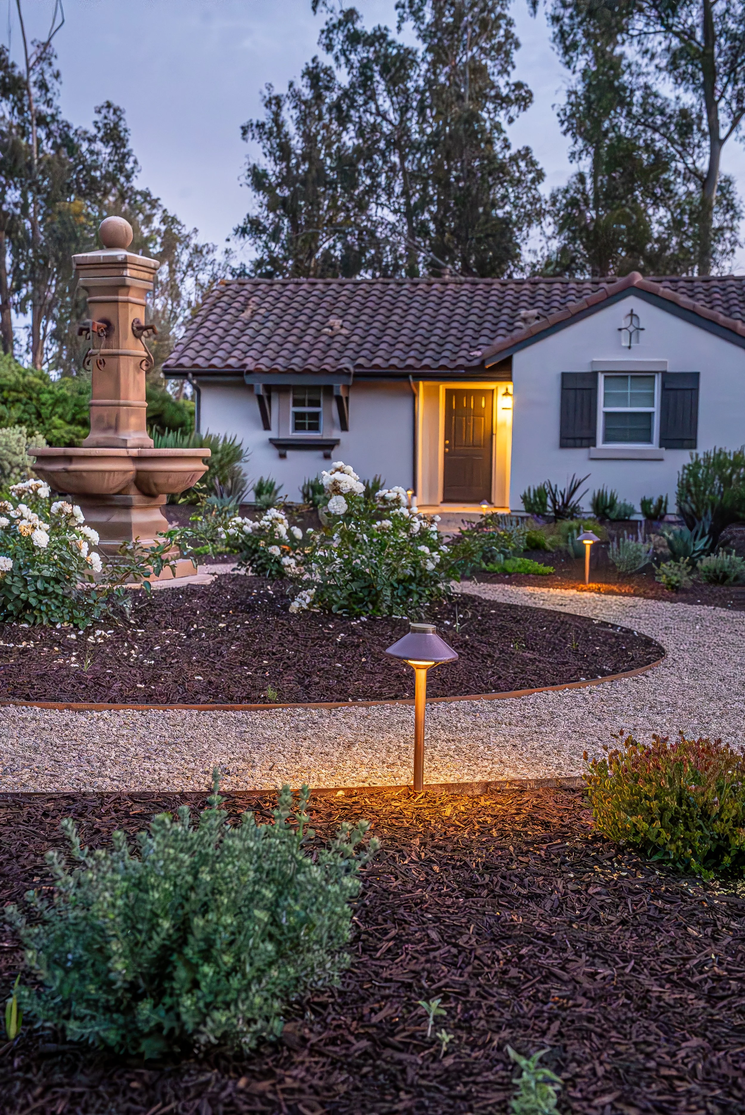 A small house with a tile roof and black shutters, illuminated warmly from inside, surrounded by a garden with white roses, pathway lights, and a decorative water fountain, during dusk.