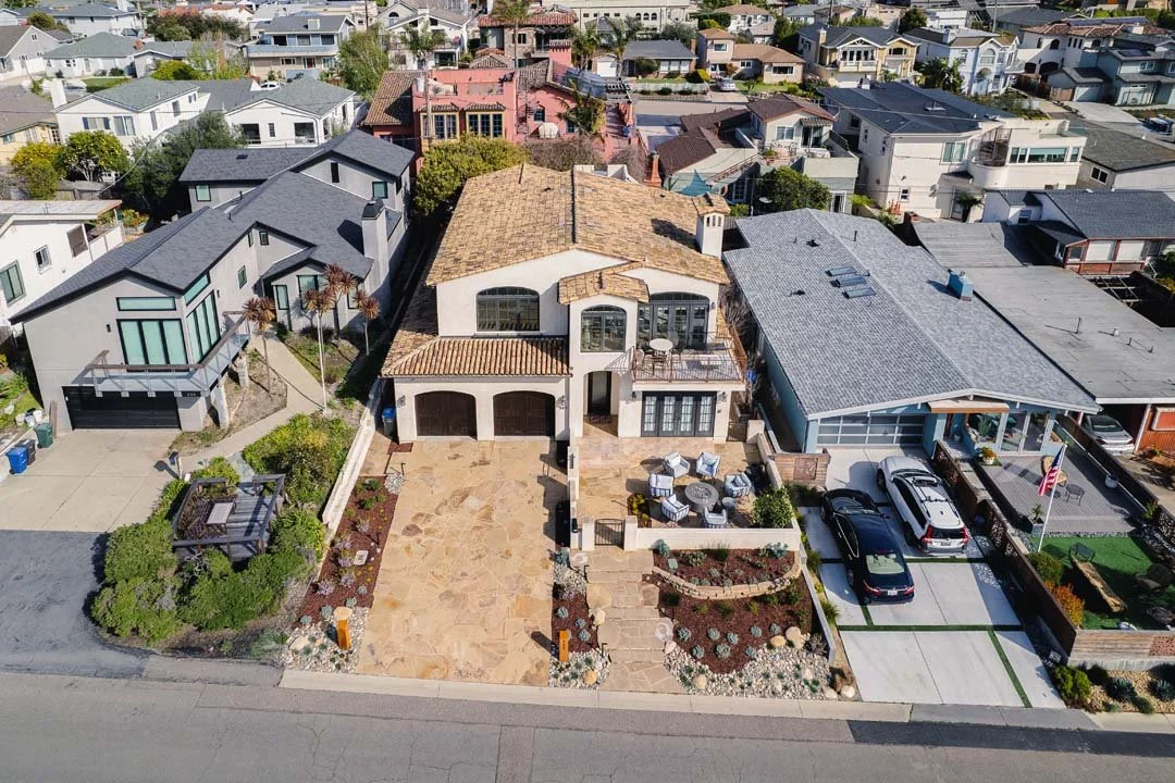 Aerial view of a suburban neighborhood with a large two-story house featuring a tiled roof, a spacious driveway with a stone patio, outdoor seating area, and a landscaped front yard designed and built by Chaparral Pavers, the top high end landscaper