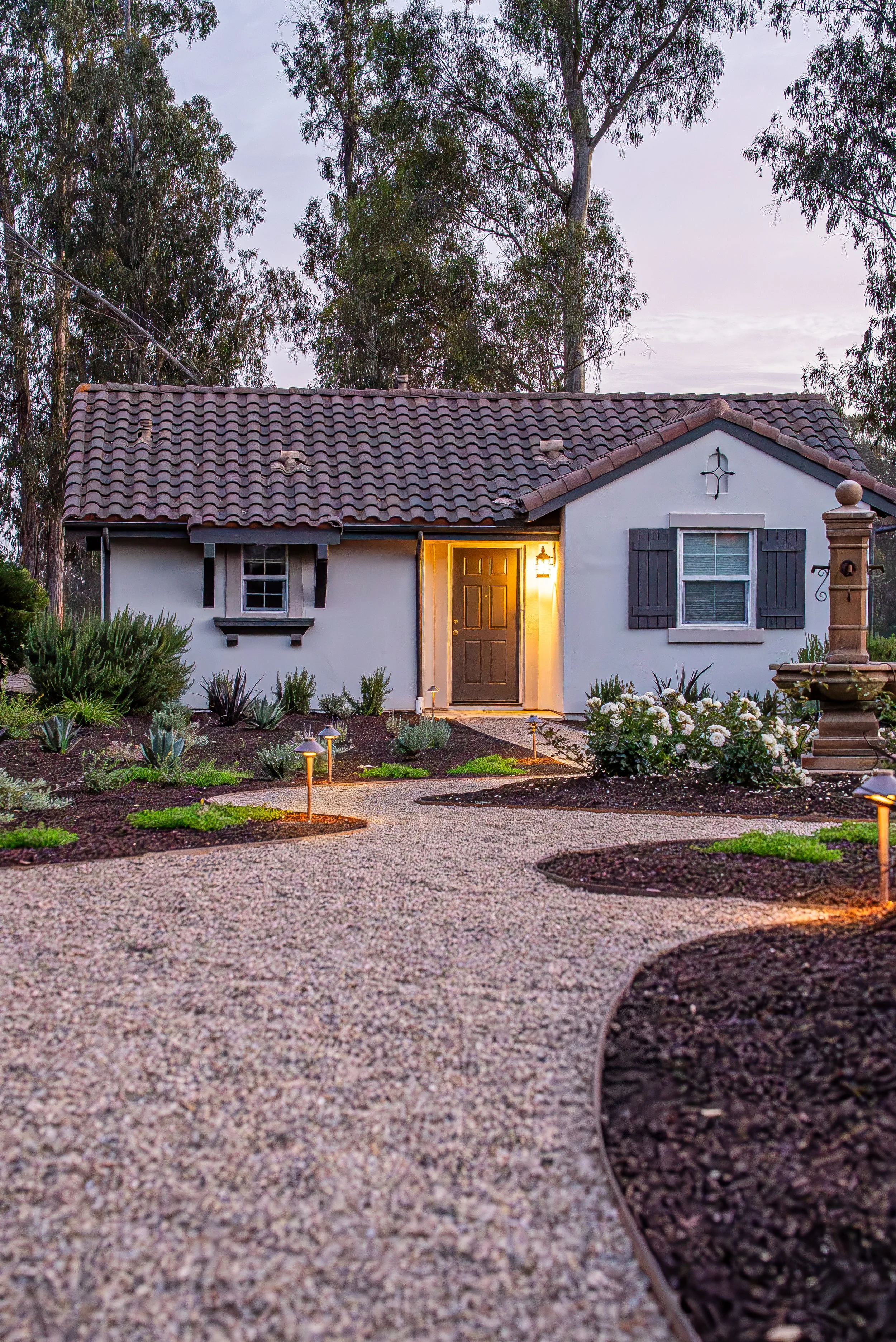 A small house with a tiled roof, white exterior walls, black shutters, and a dark front door. The front yard features a gravel pathway with garden beds and small lights, and there are numerous trees in the background. The house is lit by warm outdoor