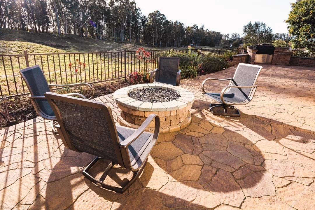 Outdoor patio with four chairs surrounding a stone fire pit, fenced yard, grassy area with trees in the background, and a brick barbecue grill on a stone patio.