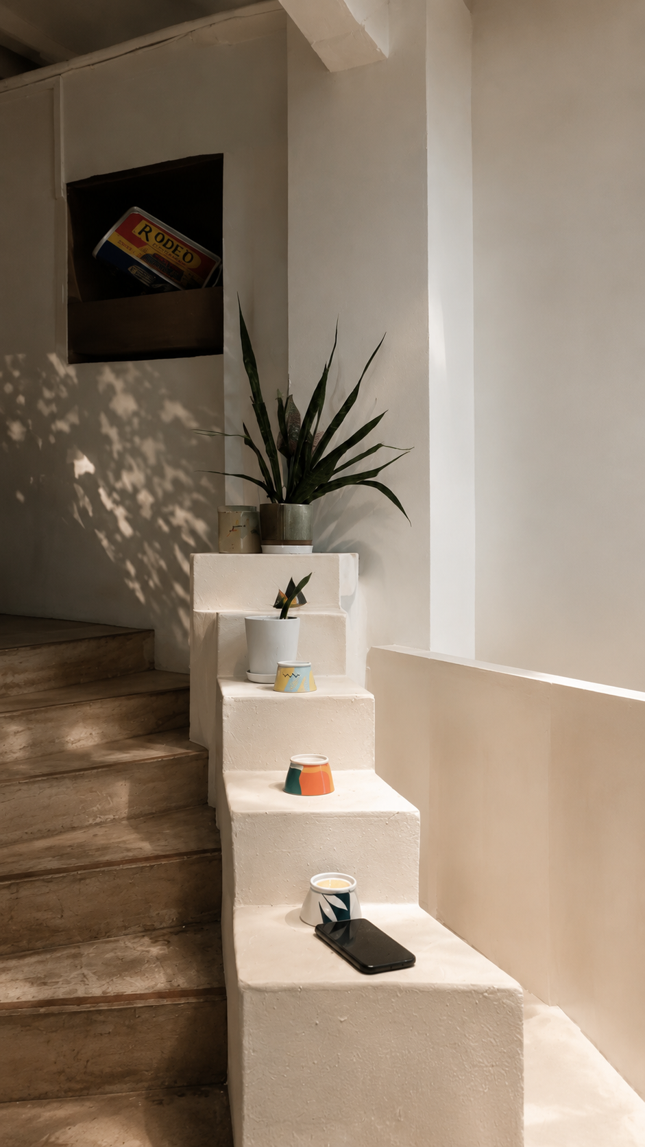 Indoor staircase with wooden steps and a white painted wall, decorated with potted plants and colorful bowls placed on the steps, with a small black object such as a phone on one of the steps.