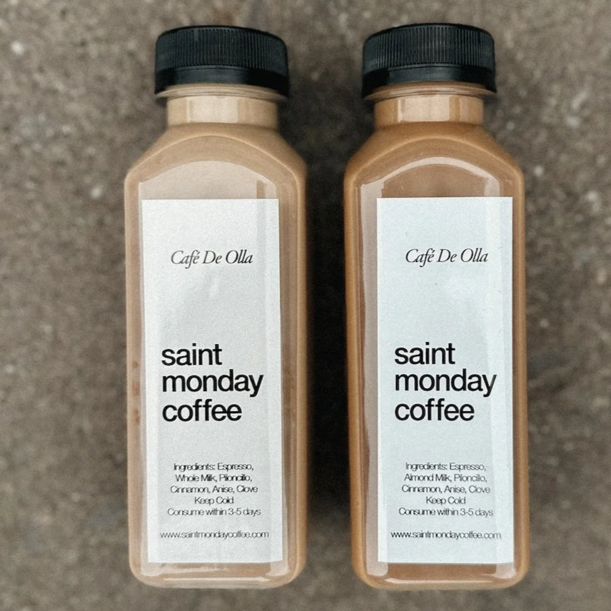 Two bottles of Cafe De Olla Saint Monday Coffee on a textured surface, with labels listing ingredients such as espresso, almond milk, plancio, cinnamon, anise, and clove.