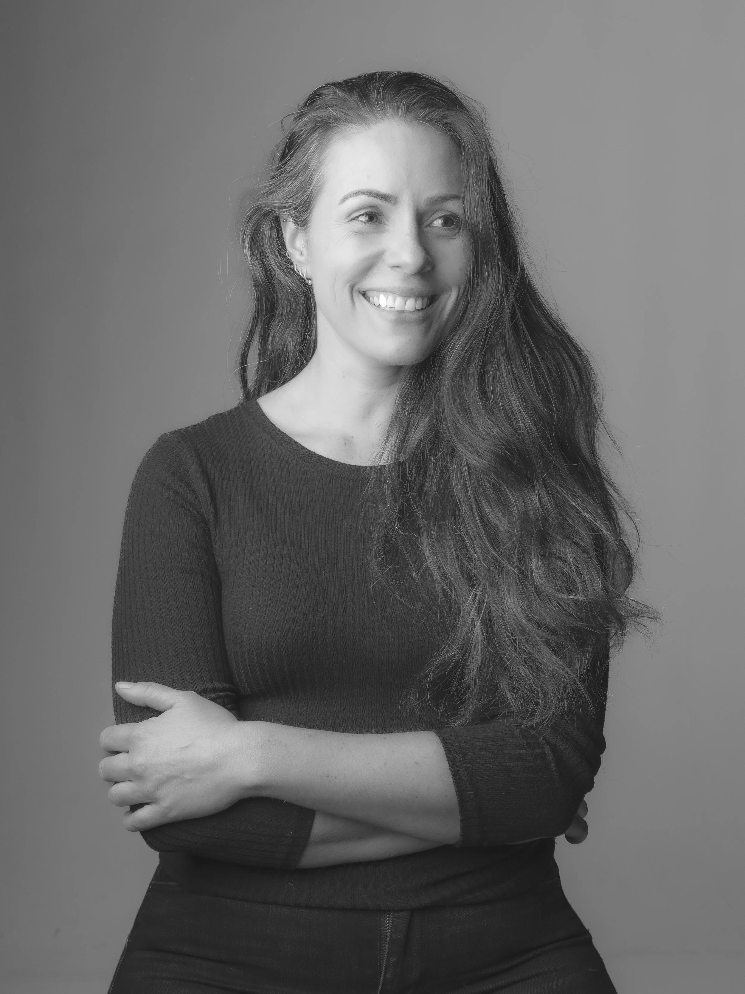 Sort-hvid portrætfotografi af en smilende kvinde med langt, bølget hår, iklædt mørk langærmet top, krydsede arme og en neutral baggrund.