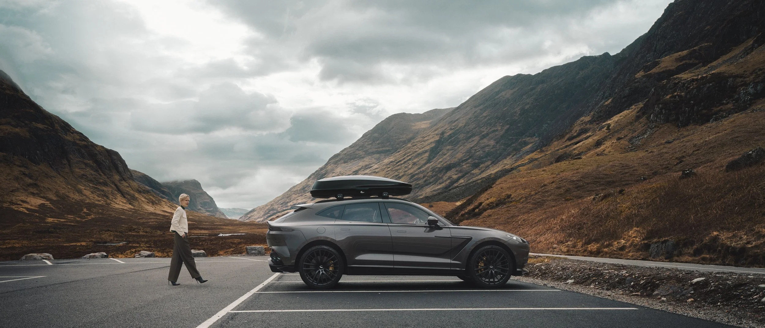 Person walking beside a luxury SUV on a quiet mountain road under stormy clouds, a cinematic automotive scene enhanced with CGI environment work and professional post-production.