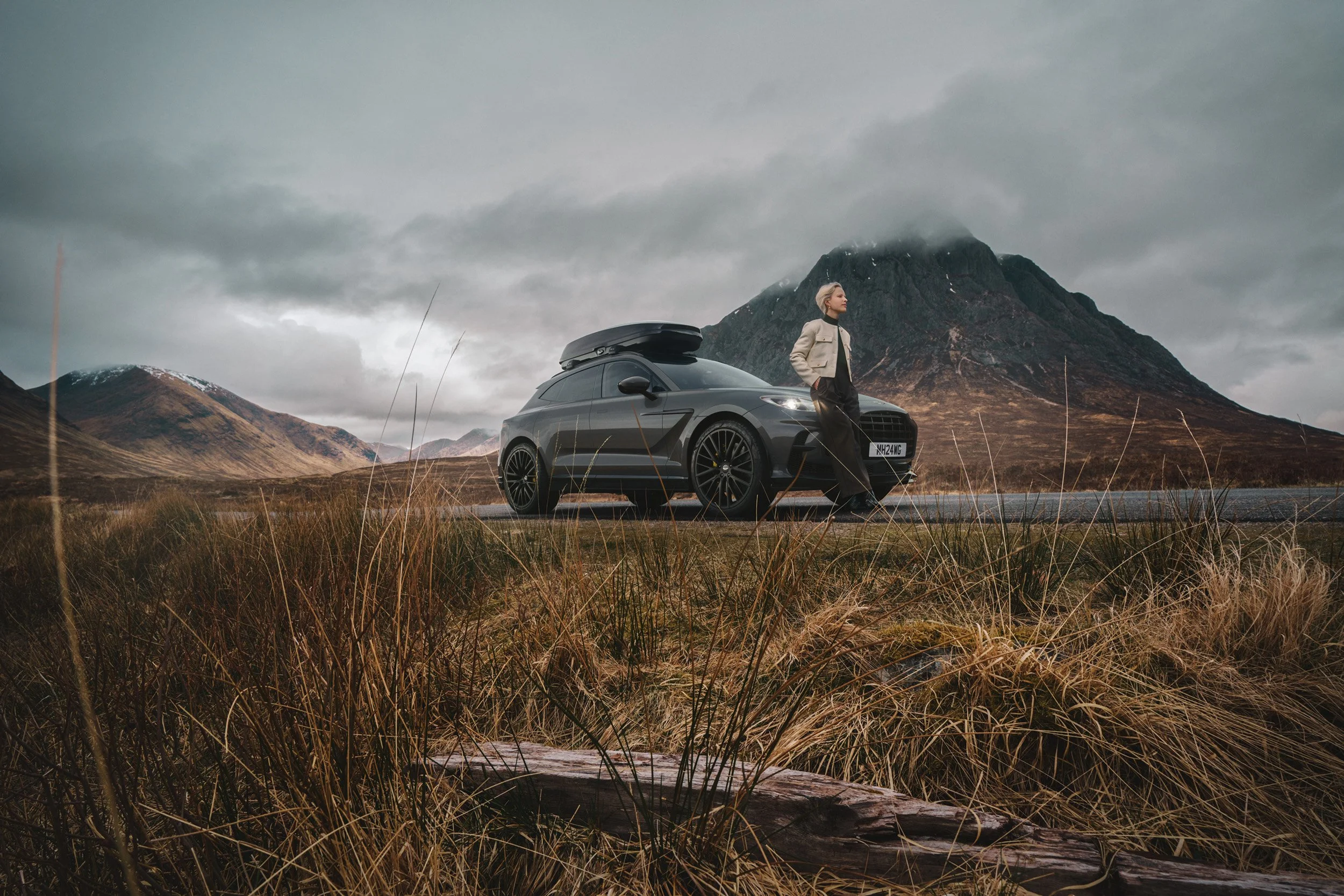 Luxury SUV parked on a remote mountain road with a person leaning against the car, dramatic cloudy highland landscape showcasing advanced CGI compositing, color grading, and post-production retouching.