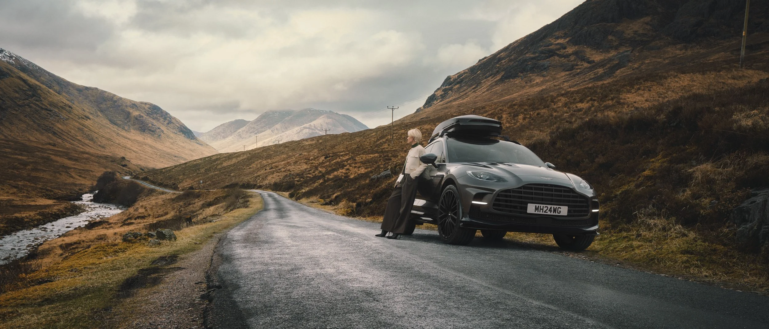 Luxury SUV parked on a dramatic mountain road with a person leaning against the car, showcasing photoreal automotive CGI compositing, retouching and cinematic post-production.