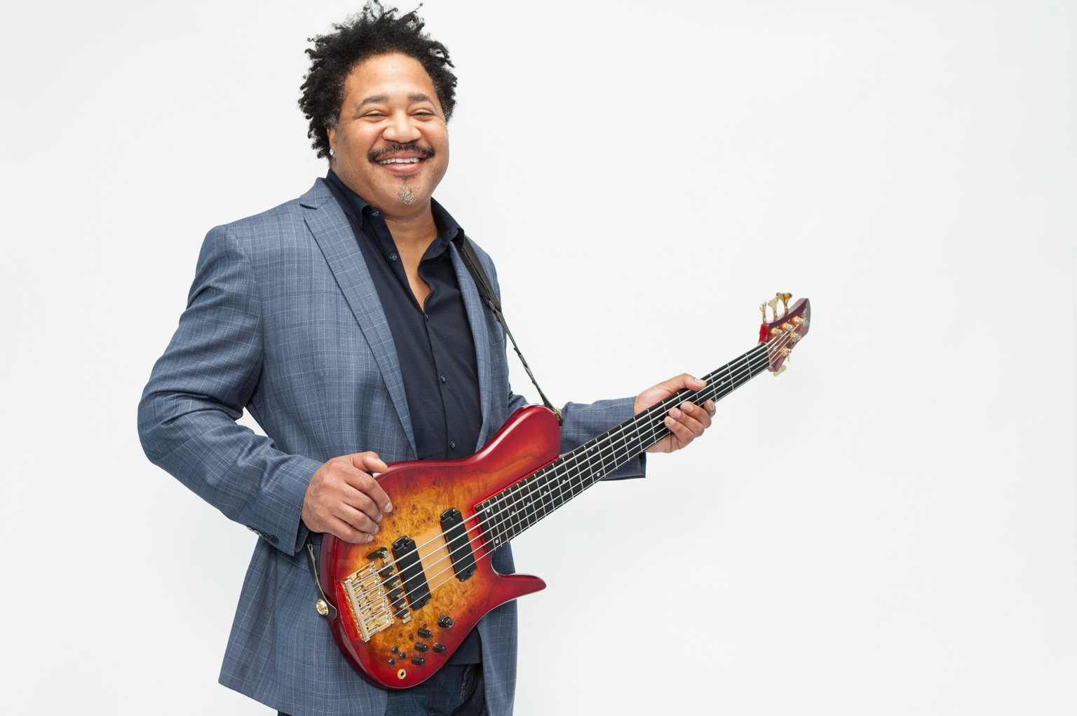 Smiling man holding a bass guitar, wearing a gray suit and black shirt, on a white background.