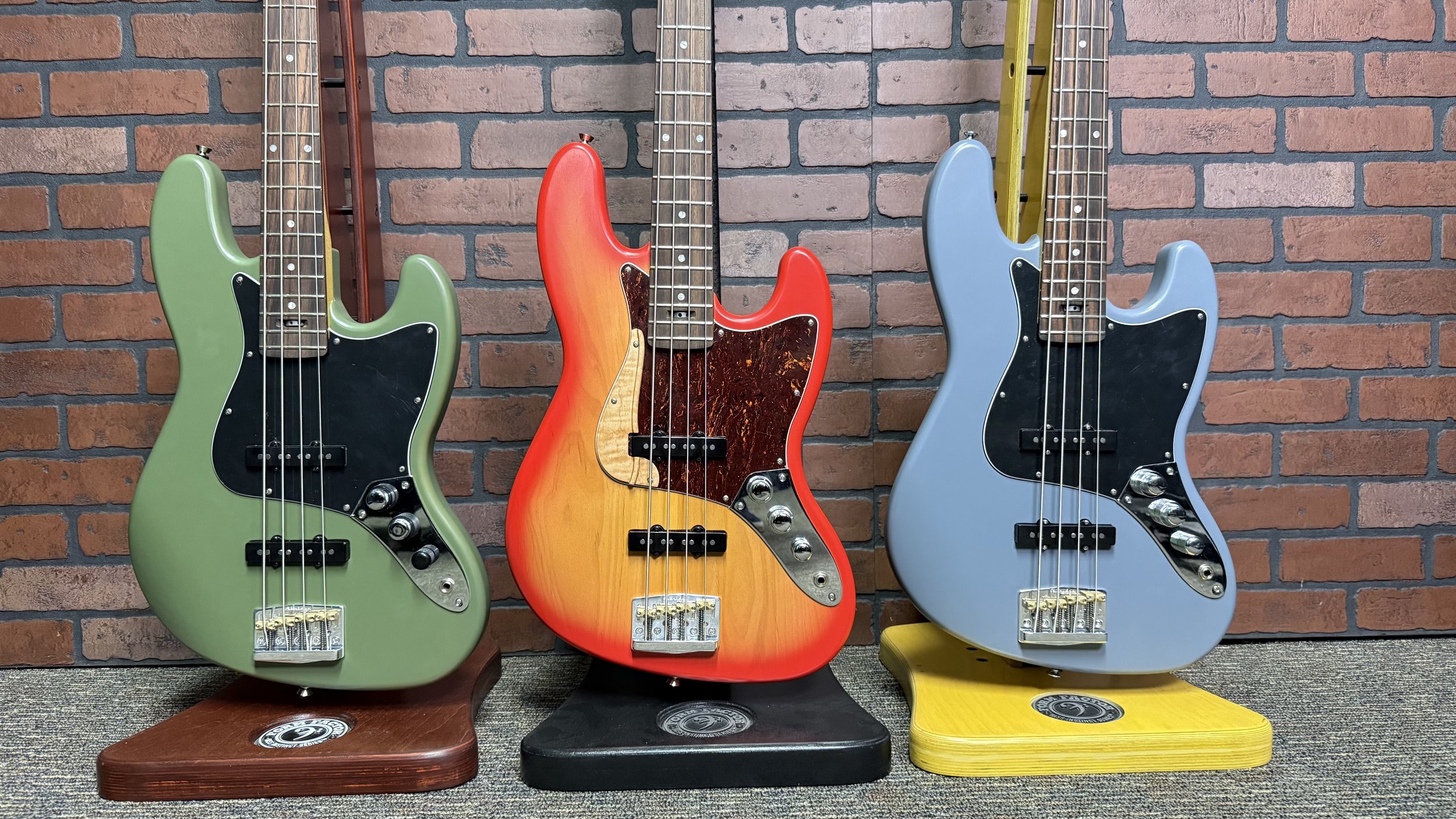 Three colorful electric bass guitars on display against a brick wall background, each on a different colored stand: green on a brown stand, red on a black stand, and blue on a yellow stand.