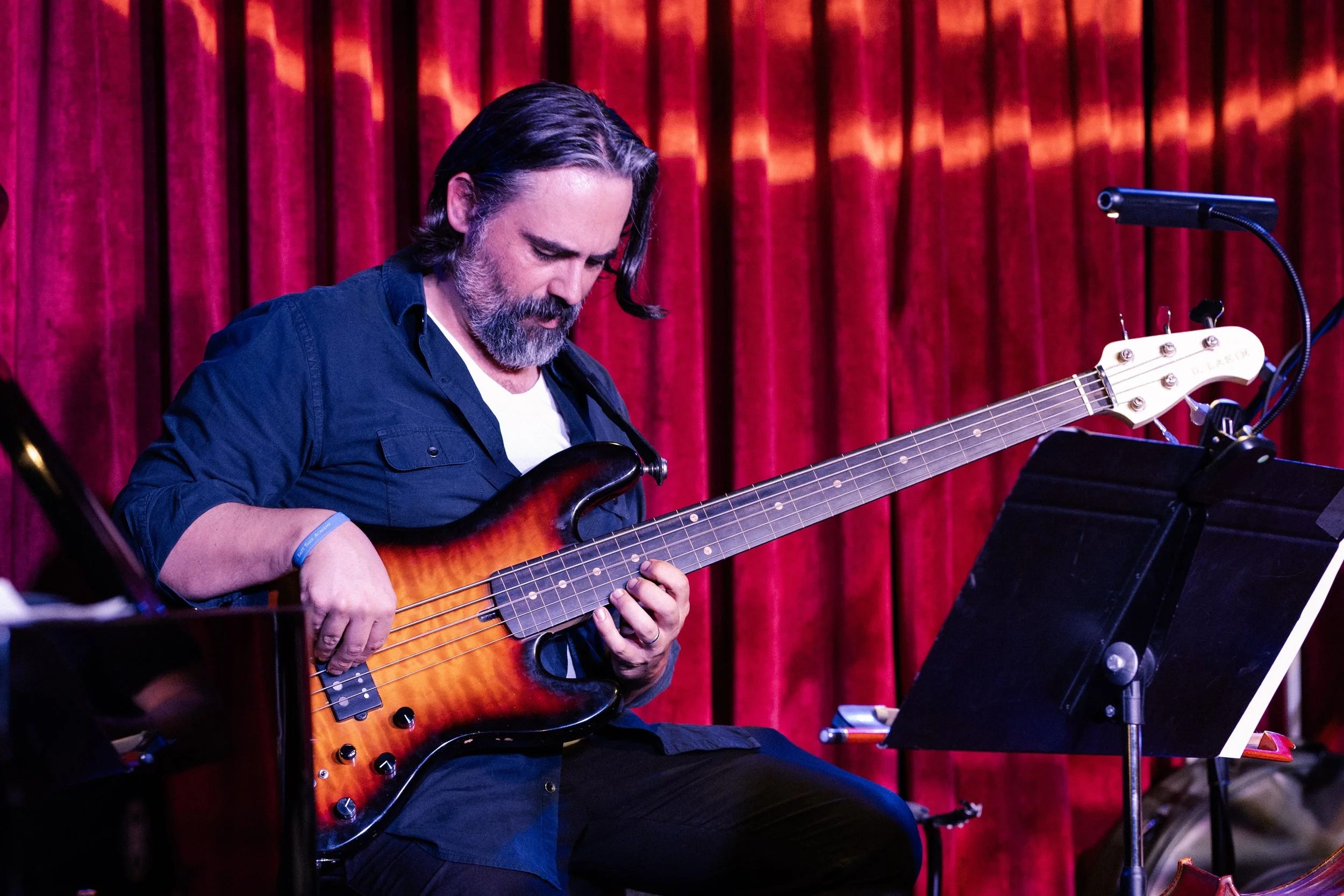 A man with long dark hair and a beard playing a six-string bass guitar with a sunburst finish on stage with a red velvet curtain in the background.