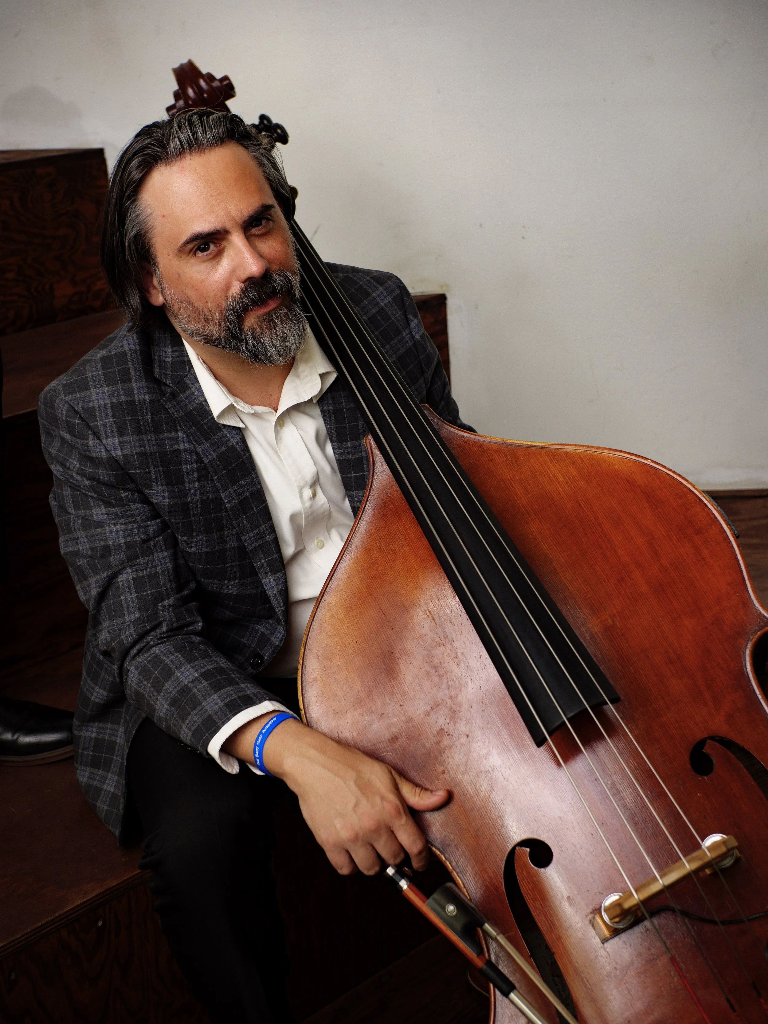 A man with gray hair and a beard, wearing a plaid blazer and white shirt, holding an upright bass, sitting in a room with a wooden floor and a white wall.